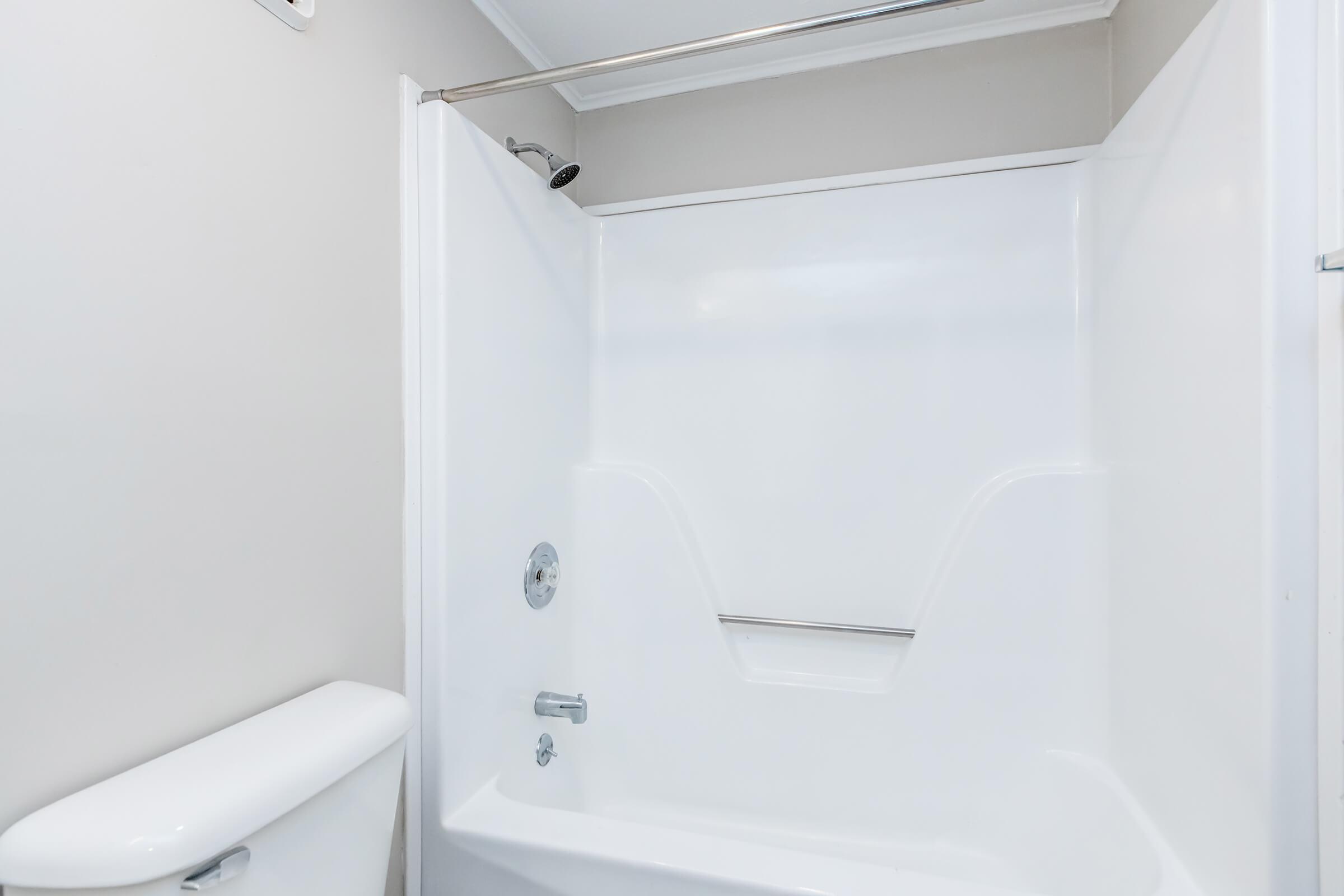 A clean bathroom featuring a white bathtub with a showerhead, a wall-mounted grab bar, and a light-colored wall. A white toilet is positioned next to the tub, creating a simple and functional space. The overall appearance is modern and well-maintained.