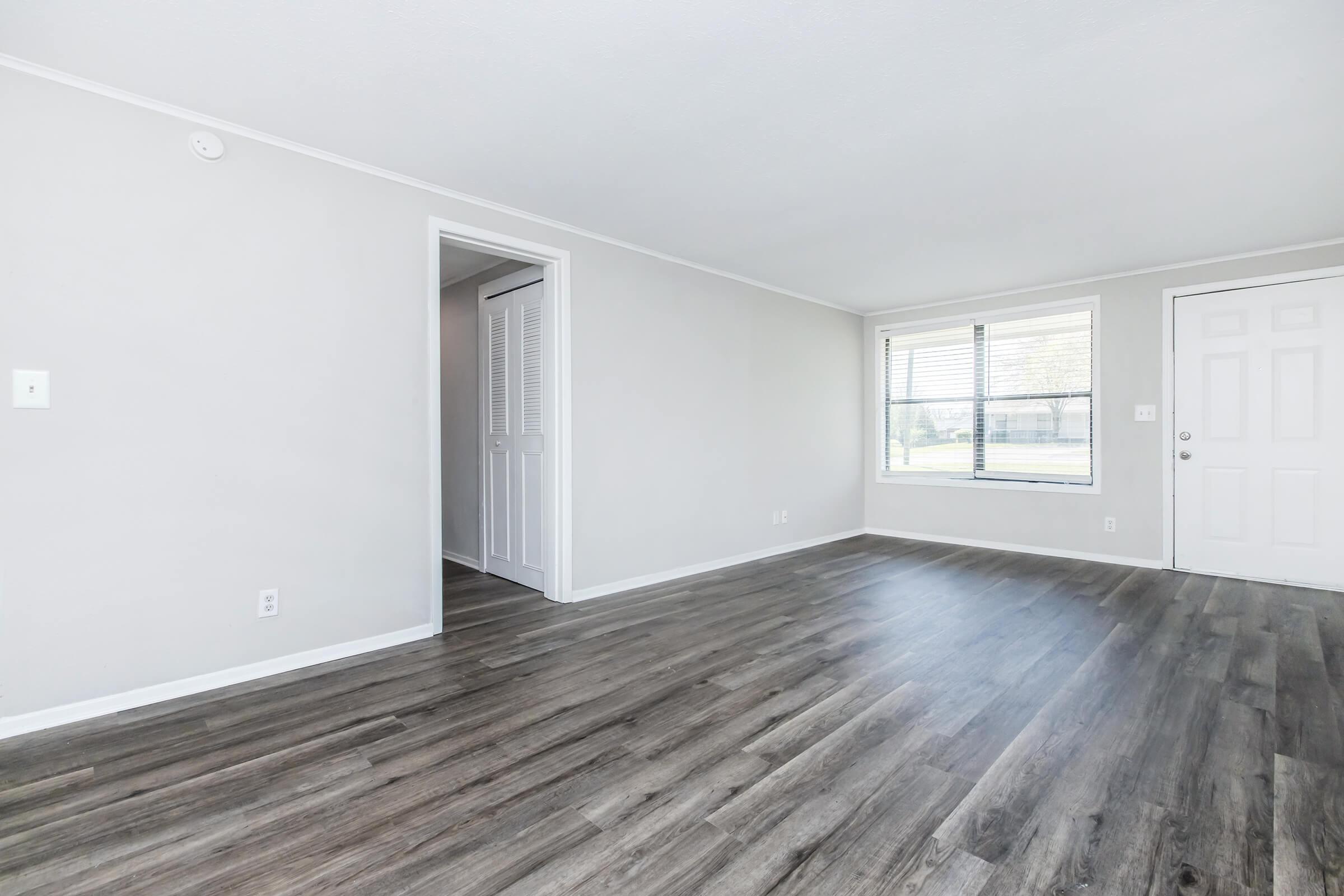 A spacious, empty interior room with light gray walls and modern flooring. There is a window with blinds allowing natural light, a doorway leading to another room, and a front door on the right. The room appears clean and well-maintained, suitable for various uses.