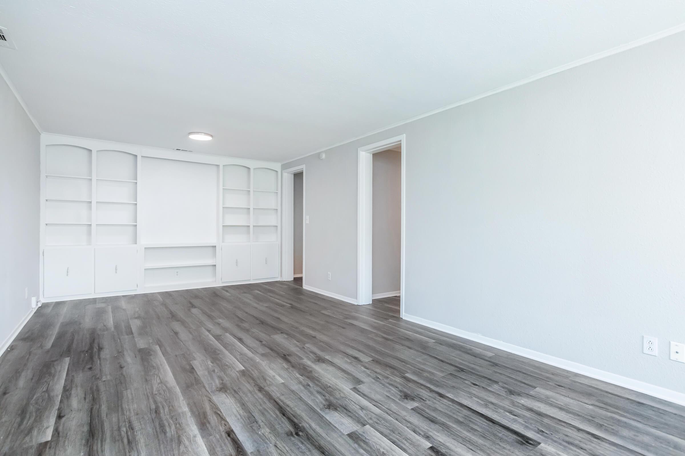 A spacious living room with light gray walls and laminate flooring. There are built-in shelves on one wall and a doorway leading to another room. The space is bright with natural light, featuring a simple, modern design.