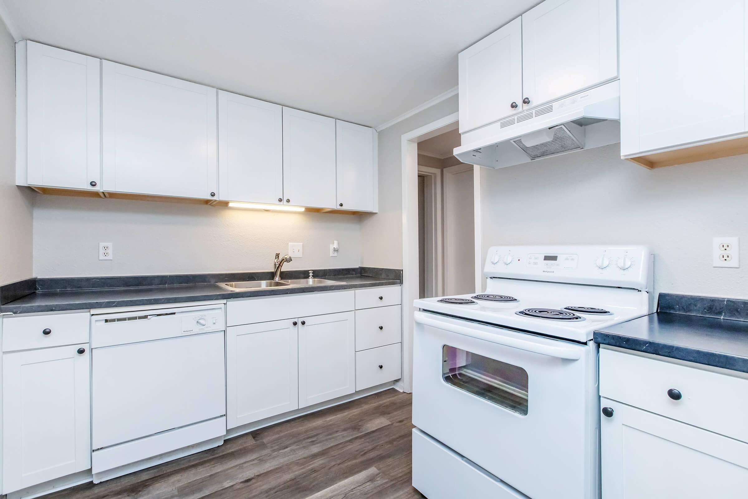 A modern kitchen featuring white cabinetry, a stainless steel sink, and black countertops. The kitchen includes a white electric stove, an oven, and a dishwasher, with hardwood-style flooring and neutral wall color, creating a clean and functional space.
