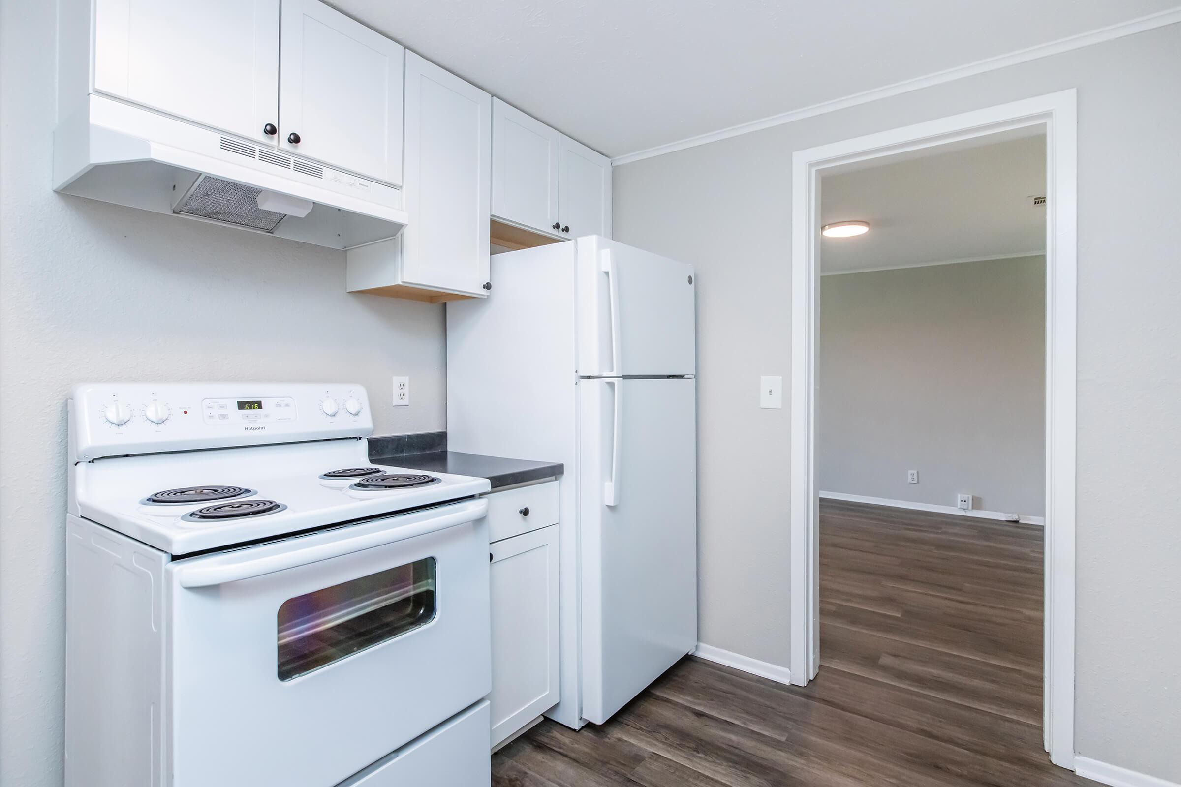 A clean, modern kitchen featuring a white stove and refrigerator, light gray walls, and laminate flooring. Cabinets are installed above the counter, creating a bright and welcoming space. An open doorway leads to an adjacent room with neutral-colored walls and flooring.