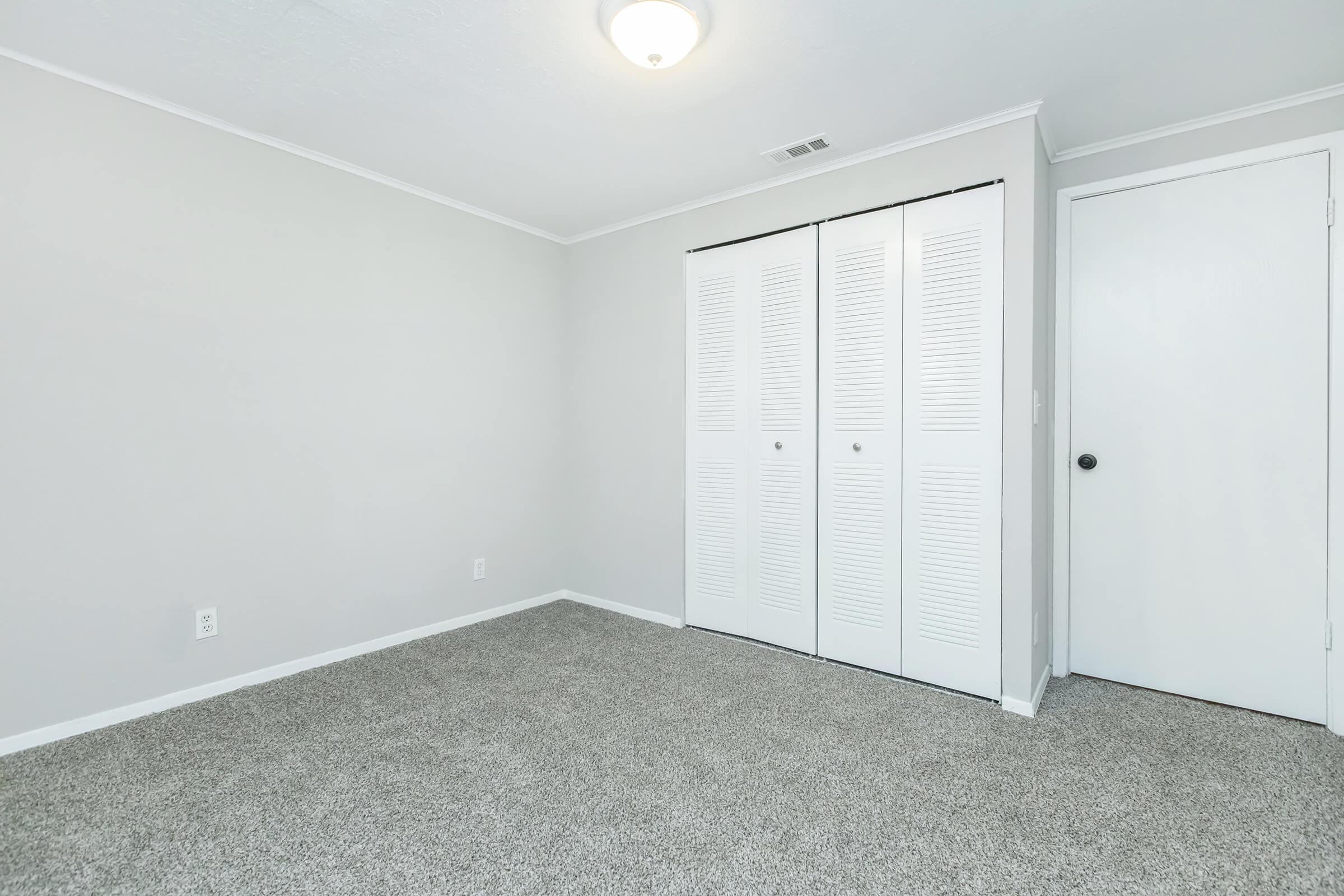 A spacious, light-colored room with beige carpet, featuring a closet with white louvered doors and a white door on the right. The walls are painted in a neutral tone and a ceiling light fixture is visible. The room appears empty and ready for furnishing.