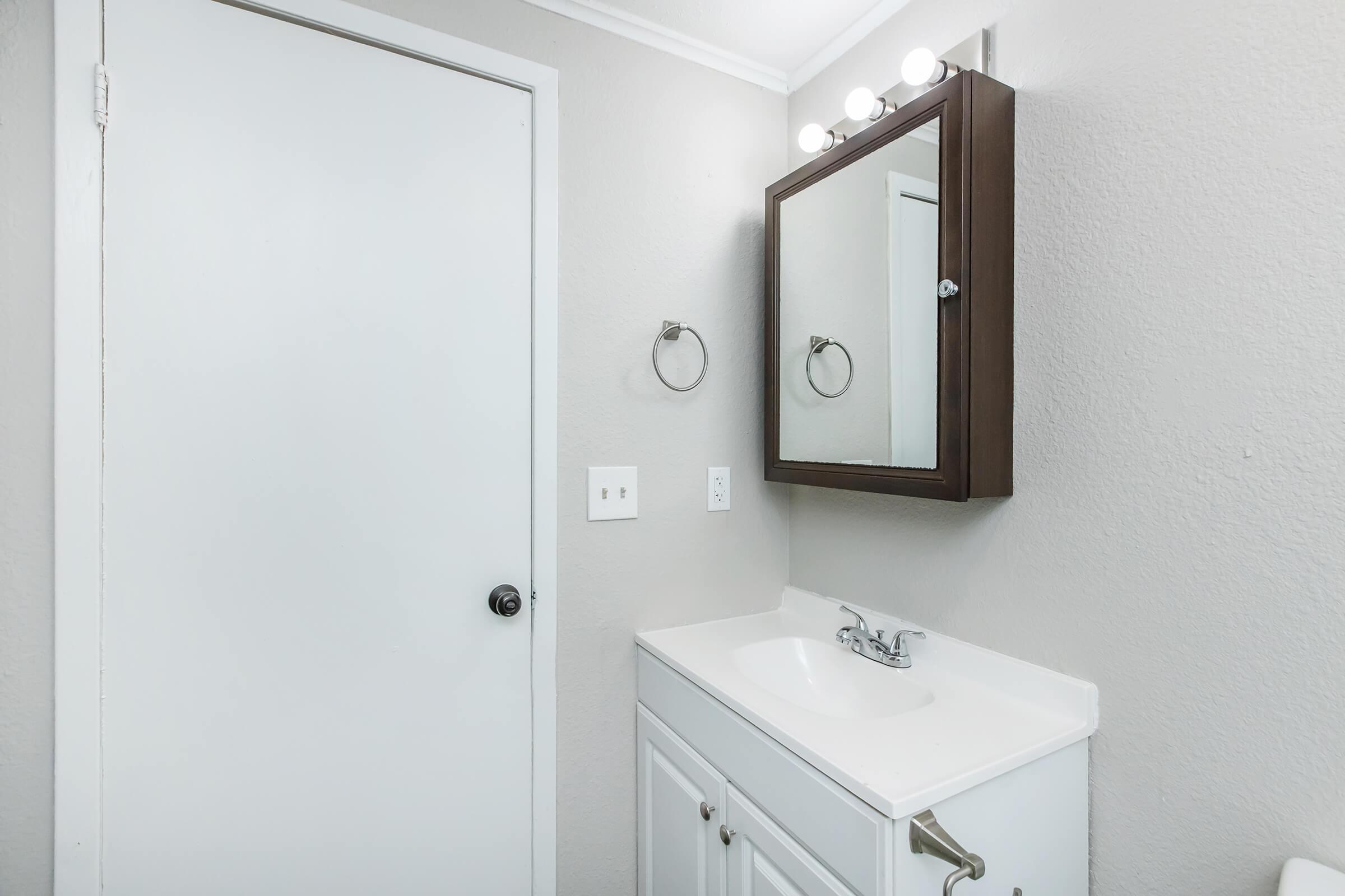 A clean and modern bathroom with light gray walls, a white vanity featuring a sink and cabinet, a rectangular mirror above the sink, and a round towel ring on the wall. A white door is visible, adding to the minimalist design. The lighting fixture above the mirror provides soft illumination.