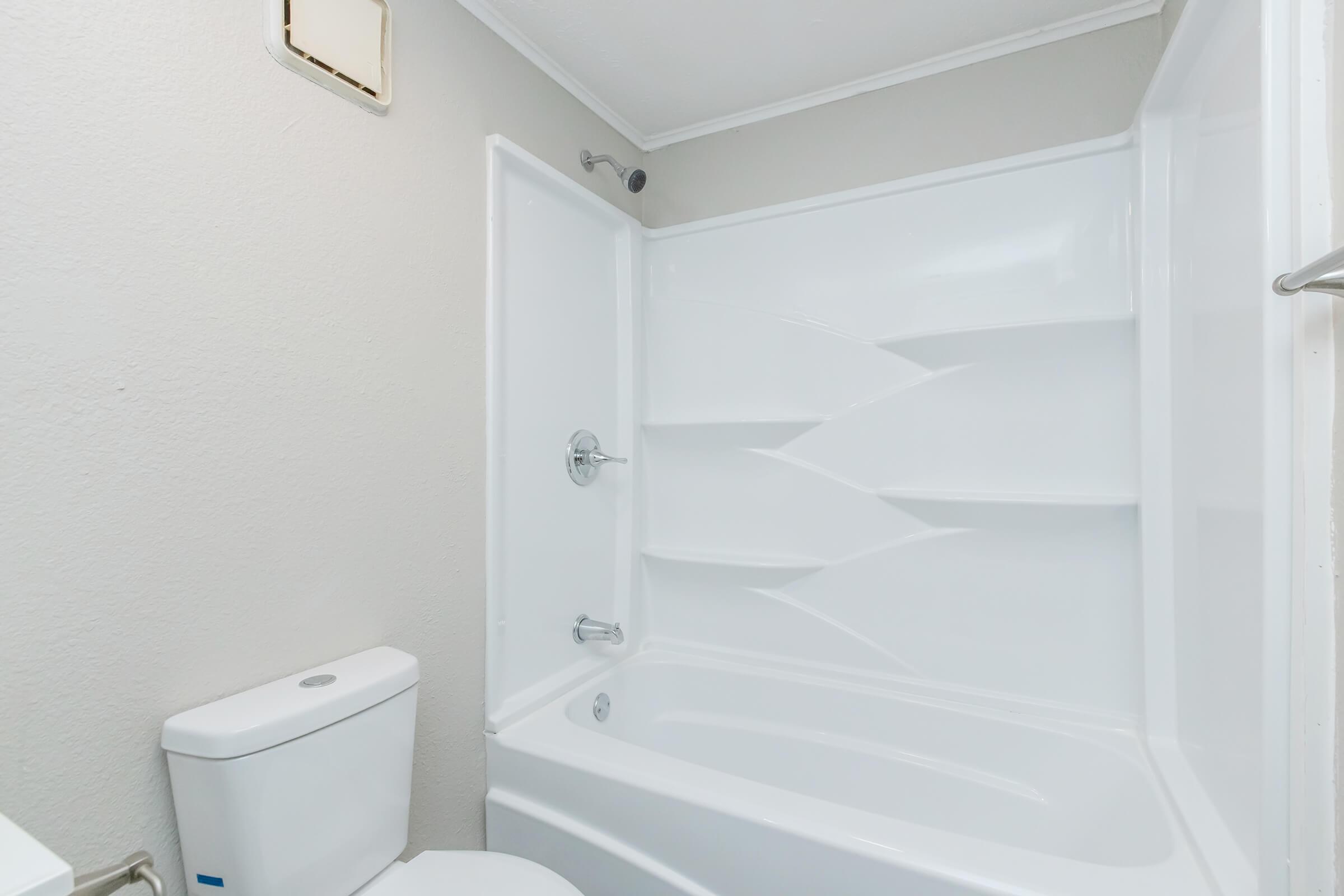 A clean, modern bathroom featuring a white bathtub and shower setup with sleek, curved wall design. There is a white toilet nearby, and the walls are painted in a light gray color, creating a bright and spacious feel.