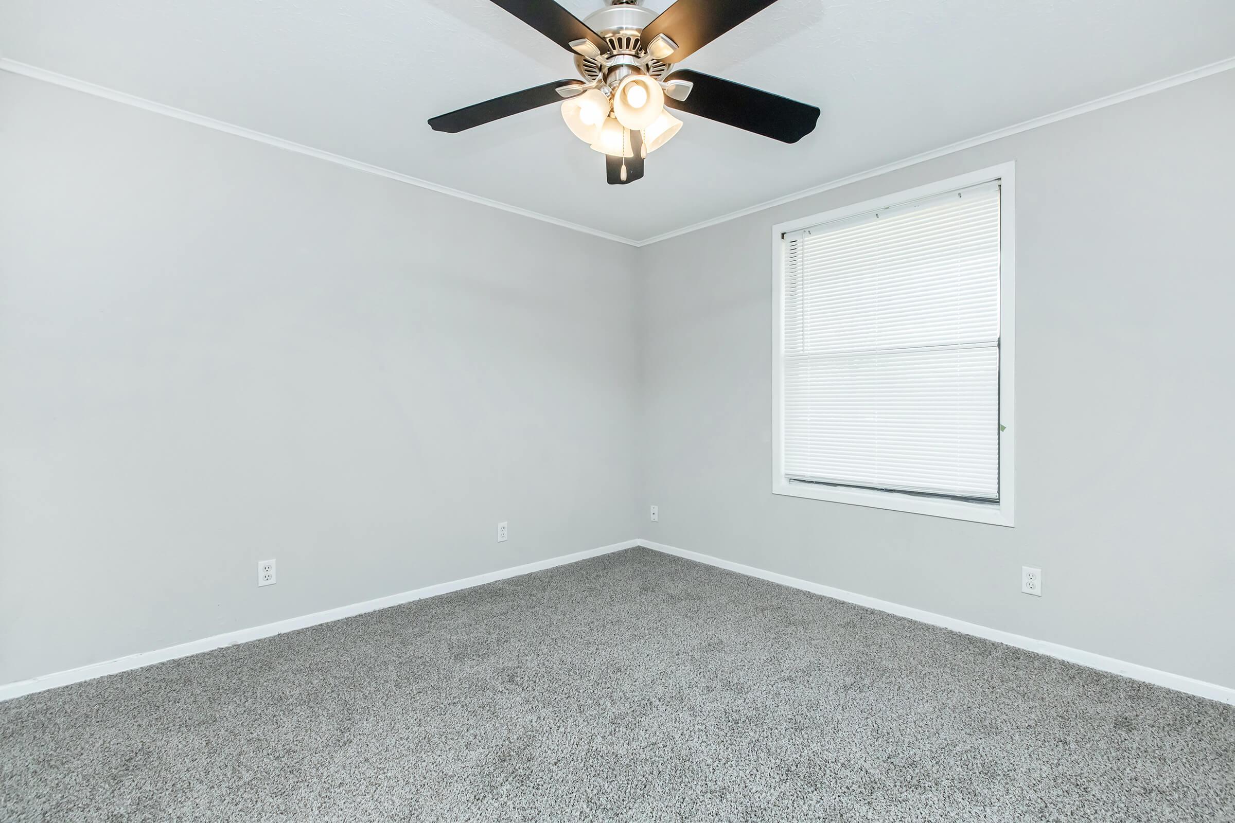 A clean, empty room with light gray walls and a soft carpeted floor. A ceiling fan with five blades is mounted above, and there is a window on the wall that allows natural light to enter. The window has horizontal blinds, providing a cozy and uncluttered atmosphere.