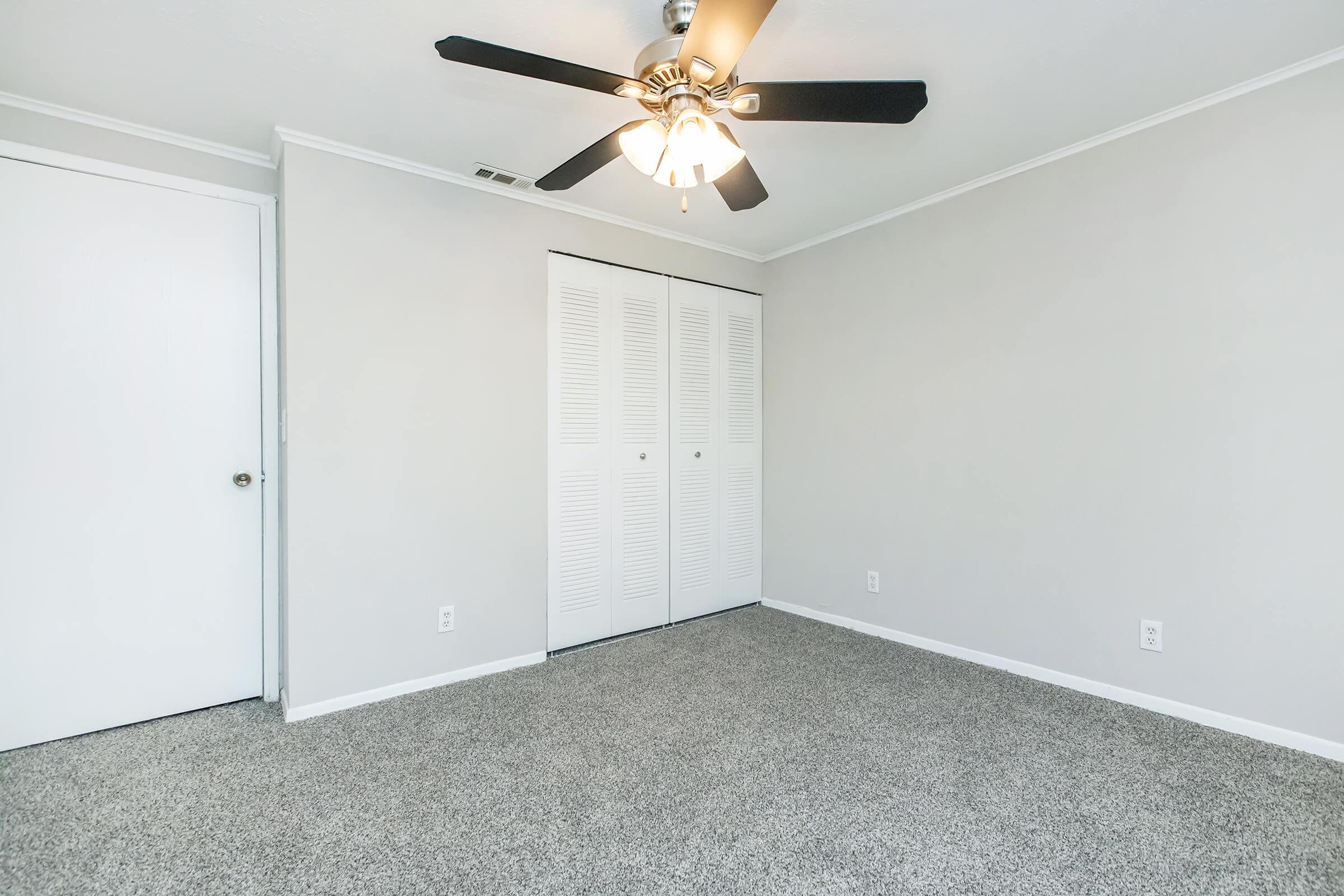 A minimalist room featuring gray carpeting, a ceiling fan with five blades, and light gray walls. There is a closed white door on the left and a closet with slatted doors on the right. The room is well-lit and empty, creating a spacious and inviting atmosphere.