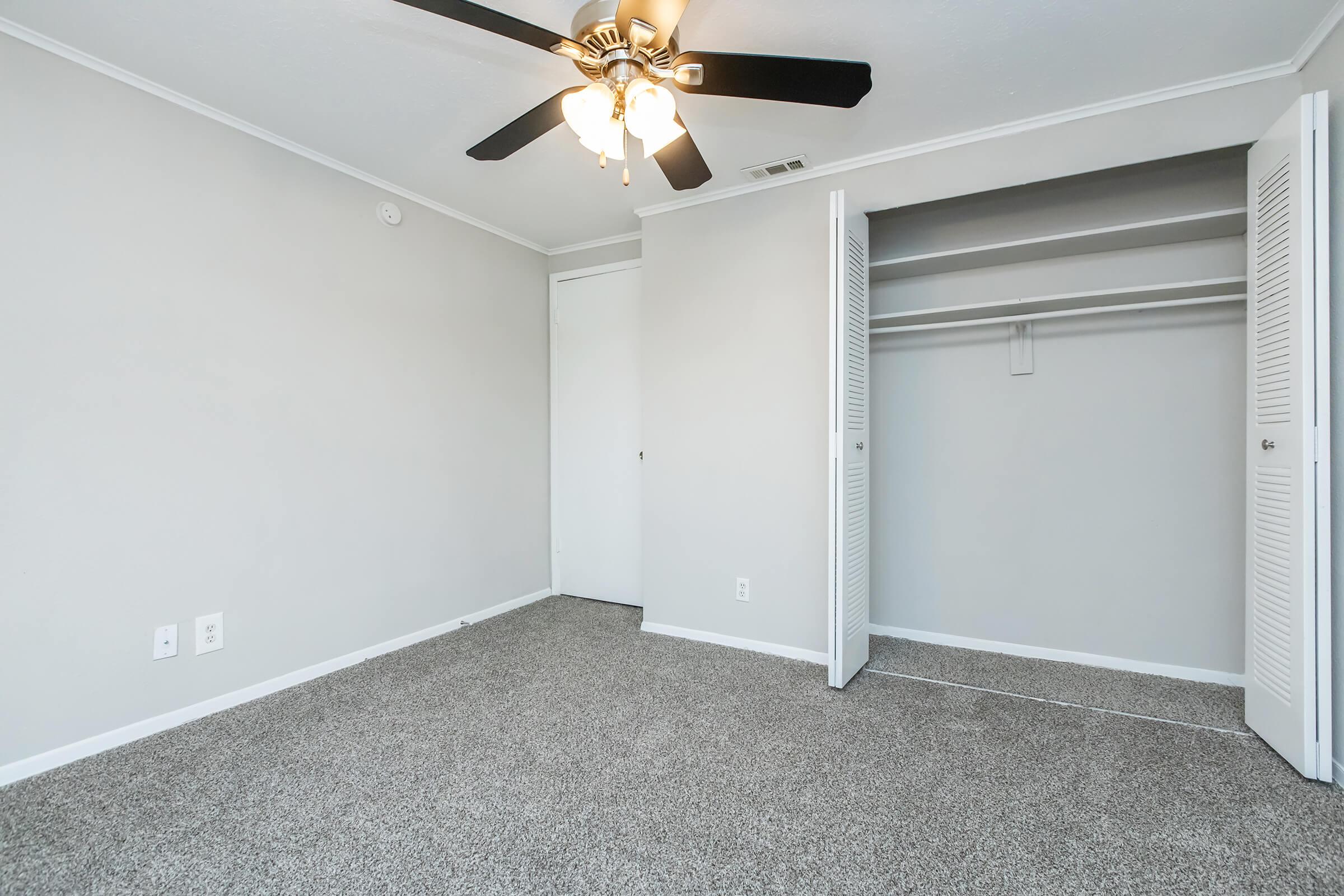A spacious, empty bedroom with light gray walls and carpet. The room features a ceiling fan with light fixtures and a closed door on one wall. There is a closet with white folding doors on the opposite wall, creating a clean and simple appearance.