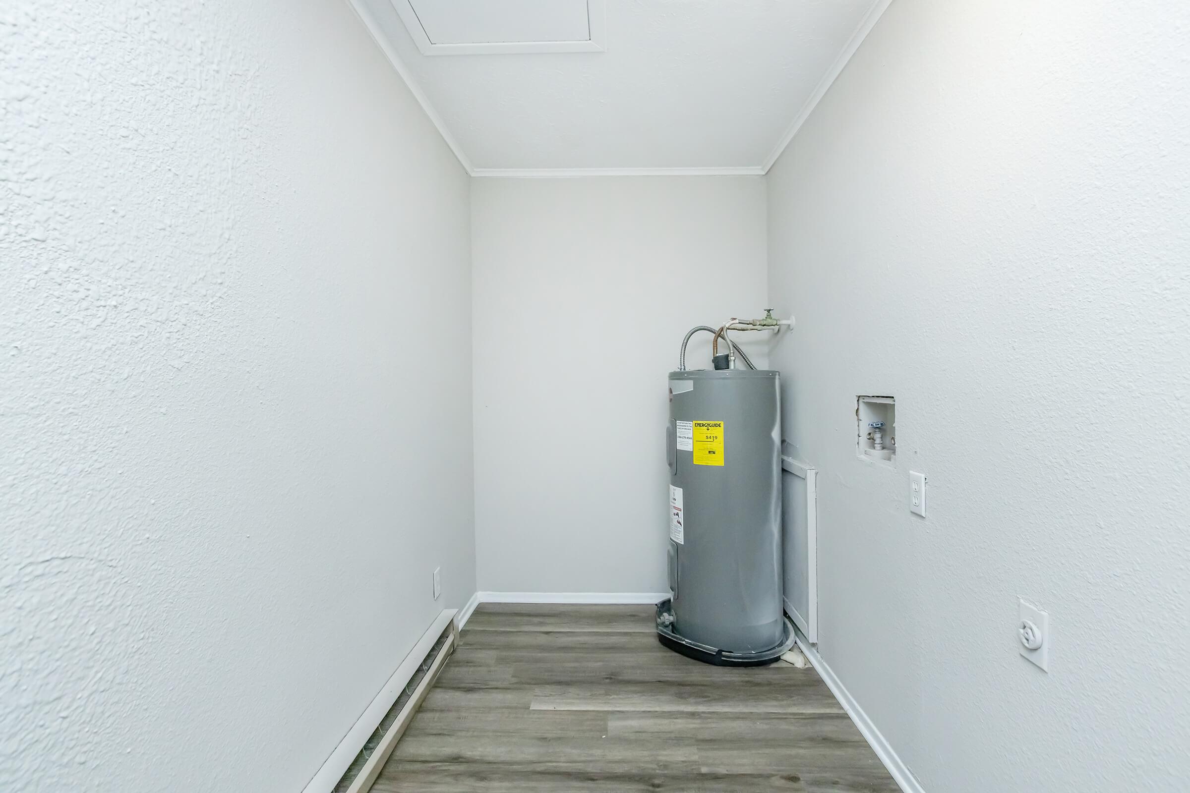 A small, empty laundry room featuring light-colored walls and a gray water heater. The space has wooden flooring and includes a wall outlet. The ceiling is white and slightly sloped, with a ceiling vent visible. The overall atmosphere is clean and simple, with ample natural light.