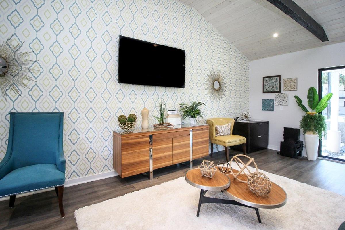 A modern living room featuring a patterned wall, a flat-screen TV mounted above a wooden sideboard, two stylish armchairs—one blue and one yellow—and decorative elements like plants and artwork. A round coffee table with decorative items sits on a light rug, adding to the contemporary aesthetic.