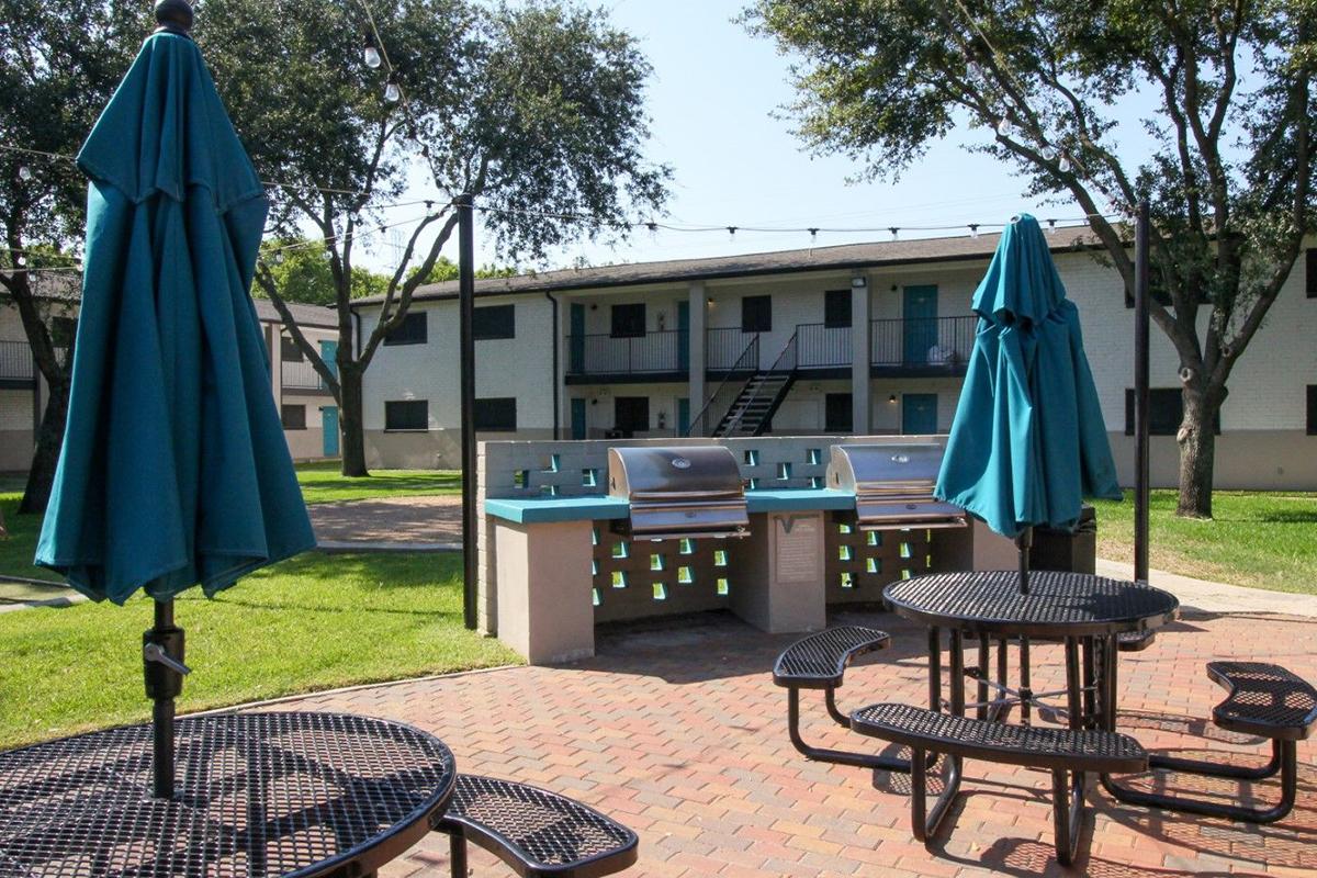 Outdoor communal area featuring picnic tables with umbrellas, a grilling station, and a green lawn. In the background, there are multiple two-story residential buildings, and trees provide shade in the area. The setting is well-lit by sunlight, creating a welcoming atmosphere.