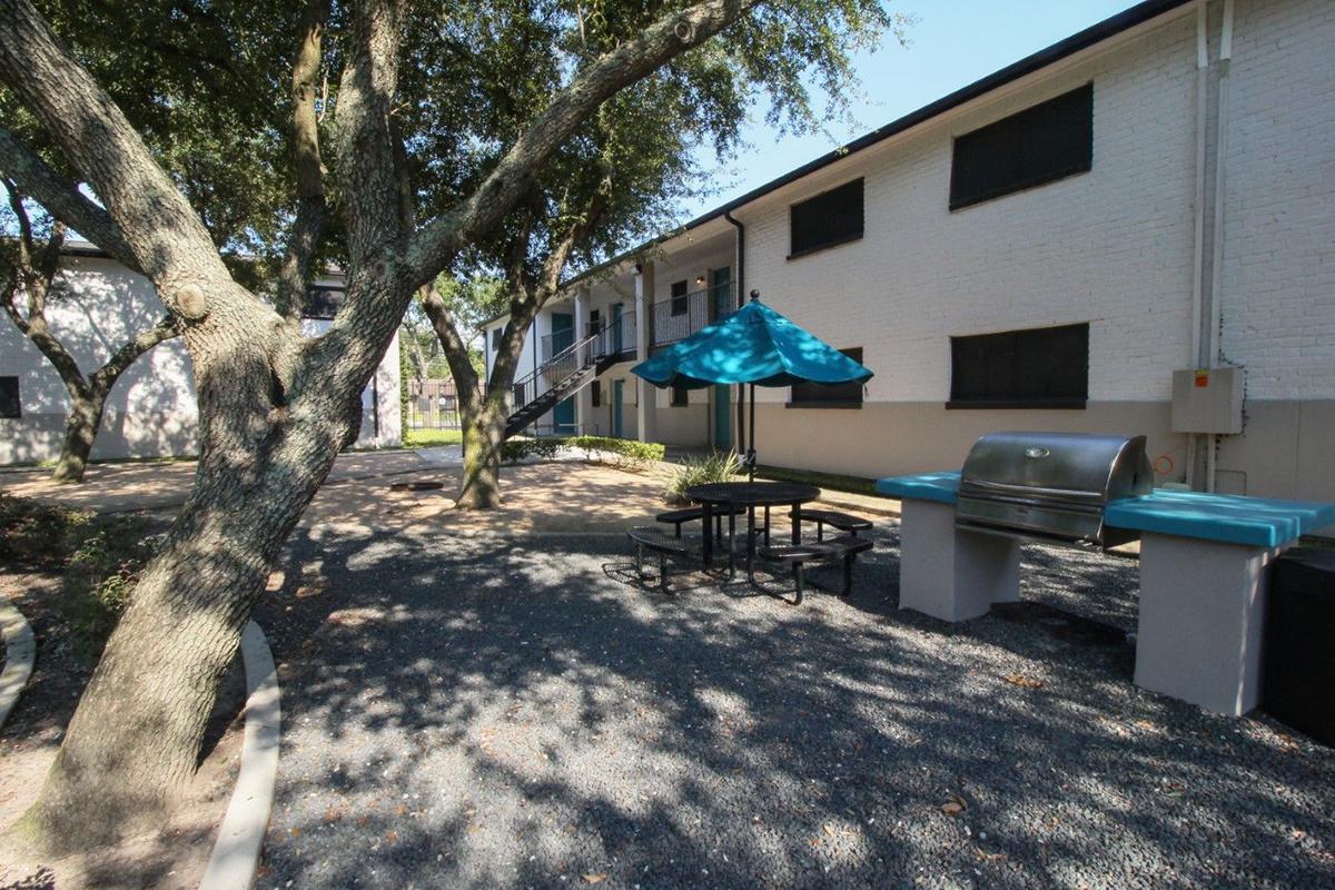 A spacious outdoor area featuring a picnic table with an umbrella, a grill, and landscaped grounds with trees. The background shows a two-story building with multiple windows and a staircase, creating a welcoming environment for gatherings or relaxation.