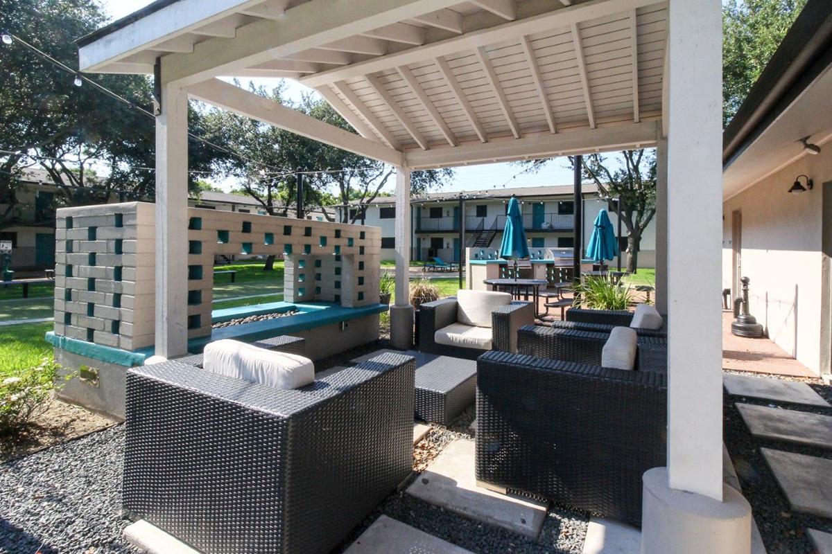 A covered outdoor seating area with modern black wicker furniture, including a sofa and chairs, placed around a small table. Surrounding greenery and a patio area with umbrellas are visible in the background, creating a welcoming atmosphere for relaxation.