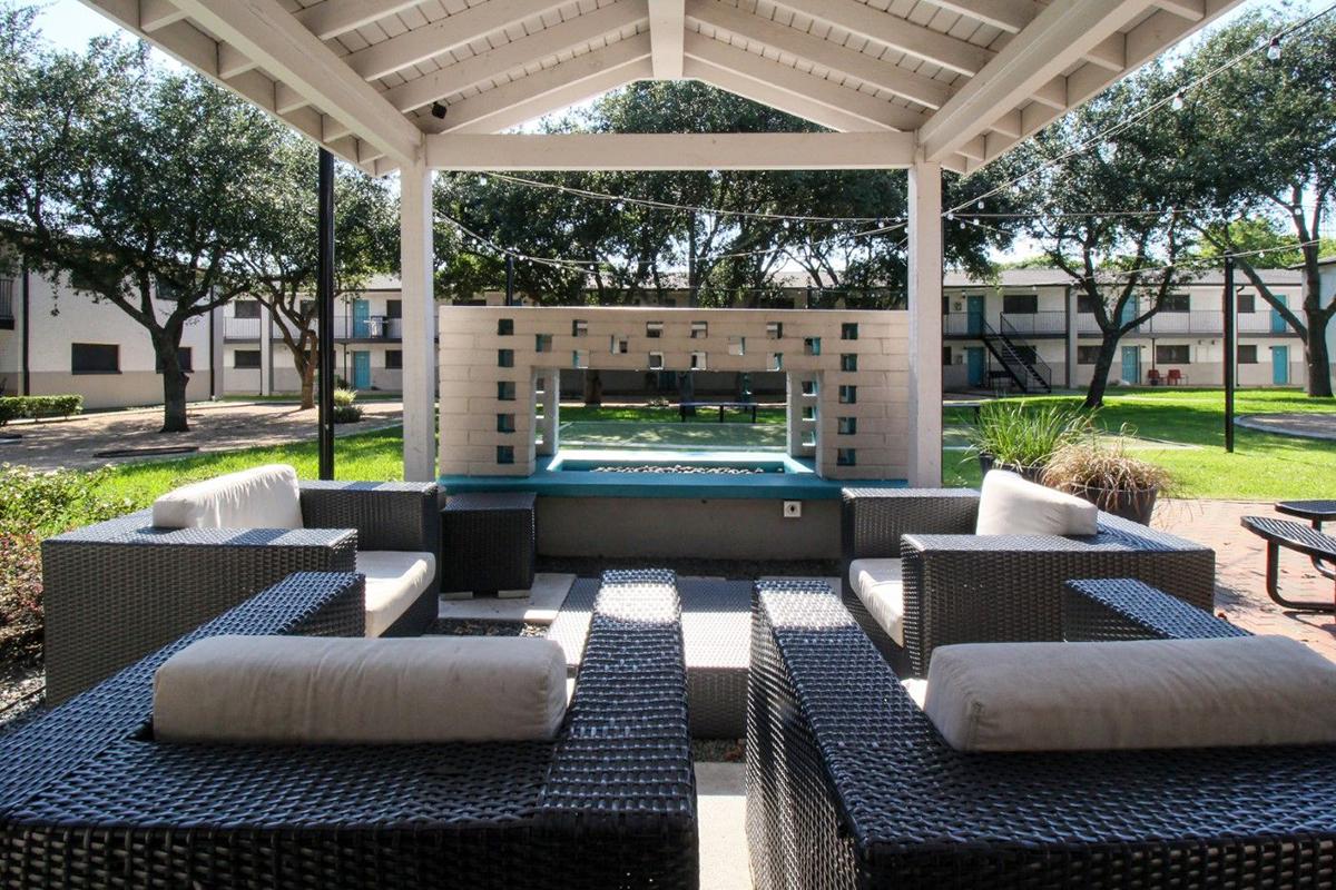 A cozy outdoor seating area with wicker chairs and cushions under a covered pavilion. The view shows a landscaped courtyard with trees and a grassy area, along with buildings in the background. Ideal for relaxation or social gatherings.