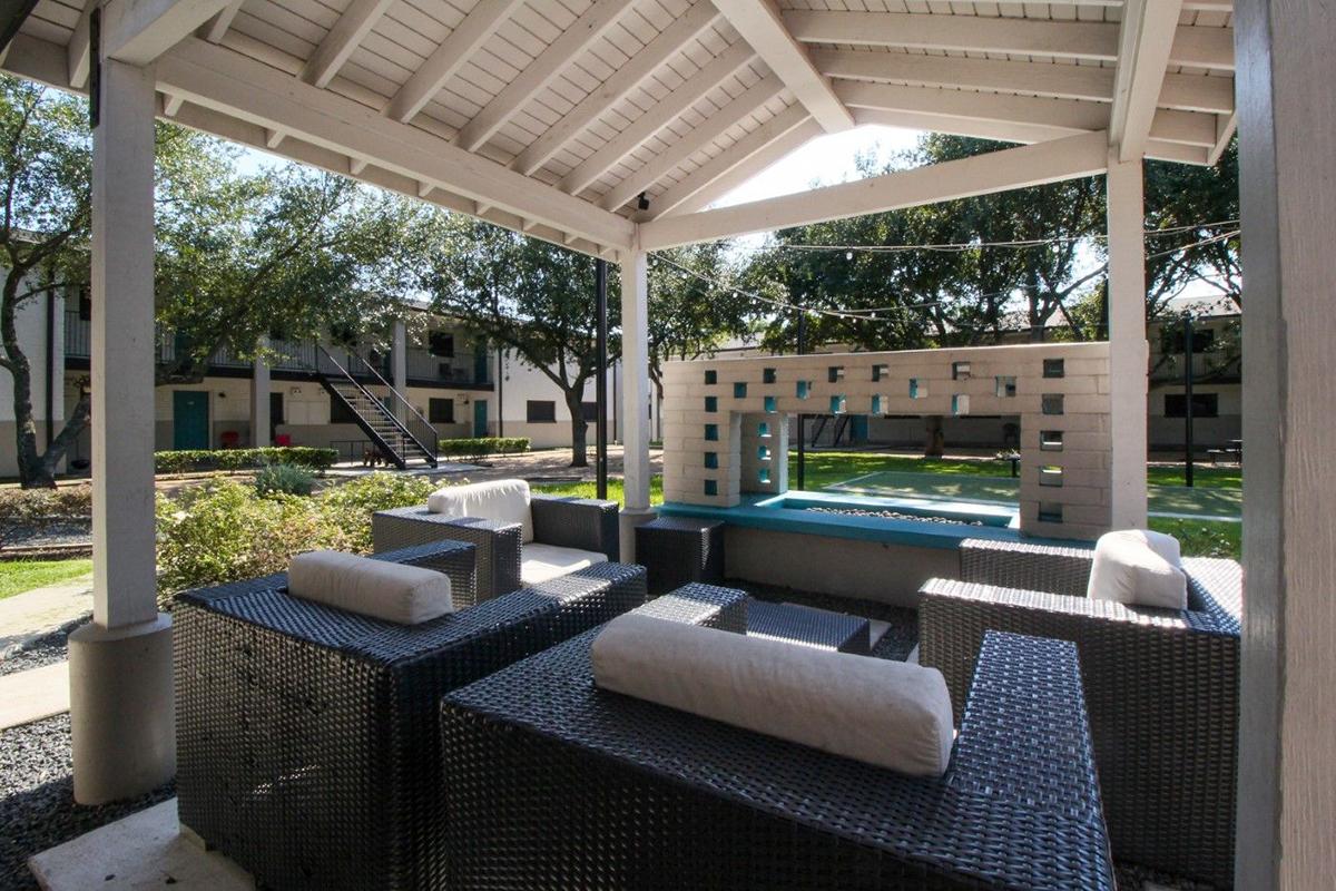 A cozy outdoor seating area featuring wicker furniture with white cushions, set under a wooden pergola. The space includes a decorative wall with a water feature, surrounded by landscaped greenery and an apartment building in the background. Perfect for relaxation and social gatherings.