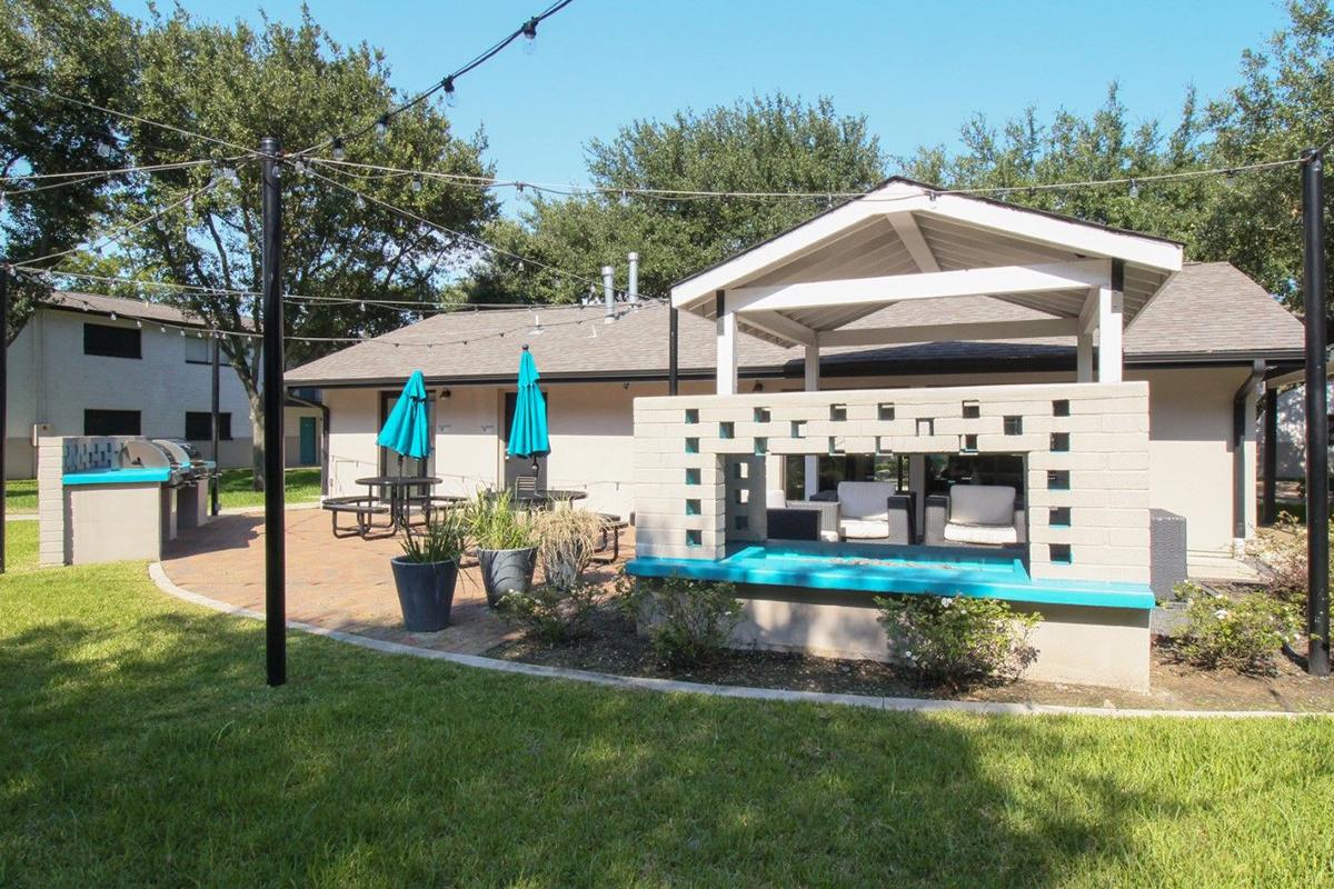 Backyard scene featuring a patio area with a covered gazebo, two teal umbrellas, and outdoor seating. There is a landscaped lawn, a small wall with a decorative cutout, and string lights overhead. The setting is bright and inviting, perfect for outdoor gatherings.