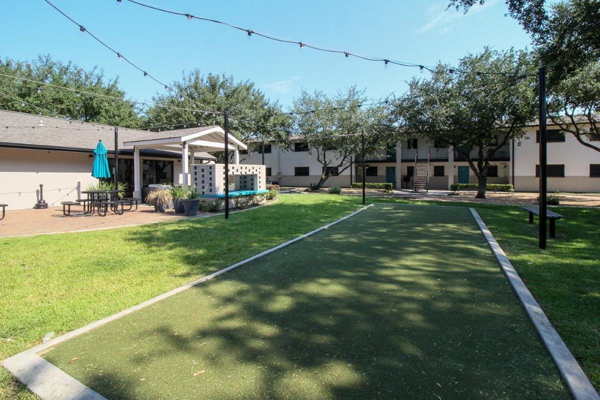 A sunny courtyard featuring a green bocce ball court, surrounded by trees and benches. There are picnic tables, a shaded area with a turquoise umbrella, and a white shelter. The space is well-maintained and inviting, with a mix of grass and paved surfaces.