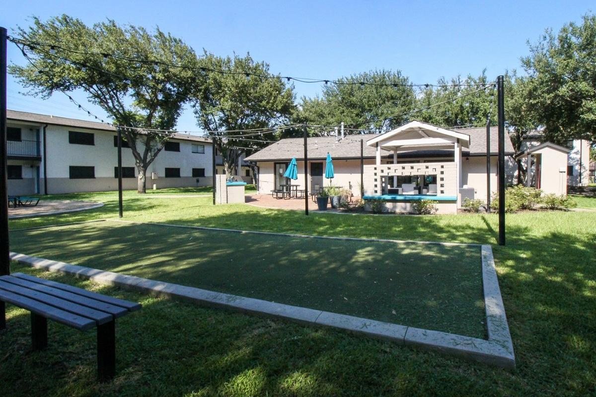 A view of a grassy area with volleyball or bocce courts, surrounded by trees and apartment buildings. There's a small pavilion with seating, and umbrellas nearby, creating a recreational space ideal for outdoor activities and relaxation.