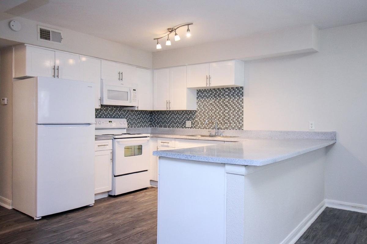 Bright, modern kitchen featuring white cabinetry, a countertop with a gray finish, a white refrigerator, stove, and microwave. The backsplash consists of decorative tiles, and the room has a light, airy feel with overhead lighting and a spacious layout.