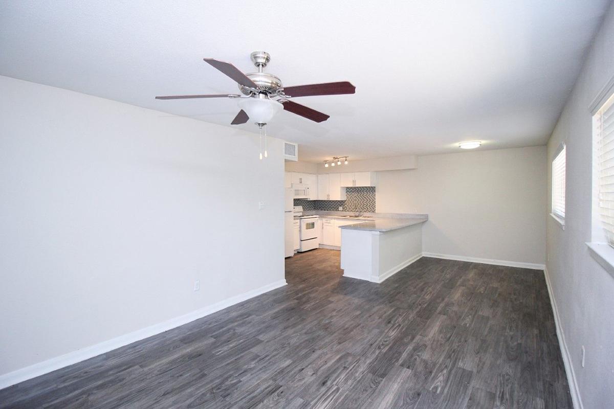 A bright and spacious living area featuring a ceiling fan, light hardwood flooring, and a modern kitchen with white cabinetry and a gray tiled backsplash. Large windows provide natural light, creating an inviting atmosphere in the room.