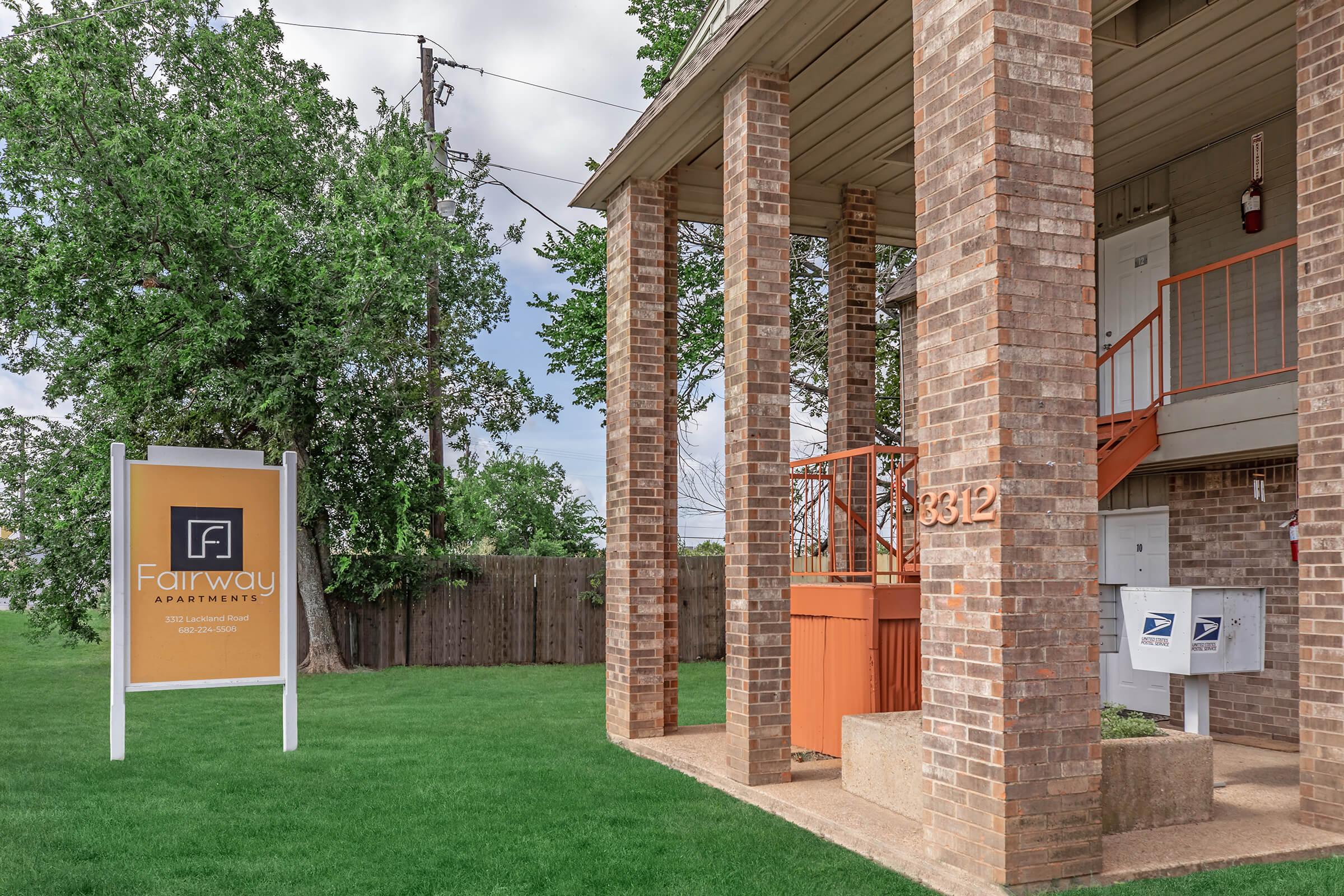 Exterior view of Fairway Apartments featuring a brick façade with four columns, a large sign promoting the apartments, and a set of mailboxes. The landscaping includes green grass and a wooden fence in the background under a partly cloudy sky.