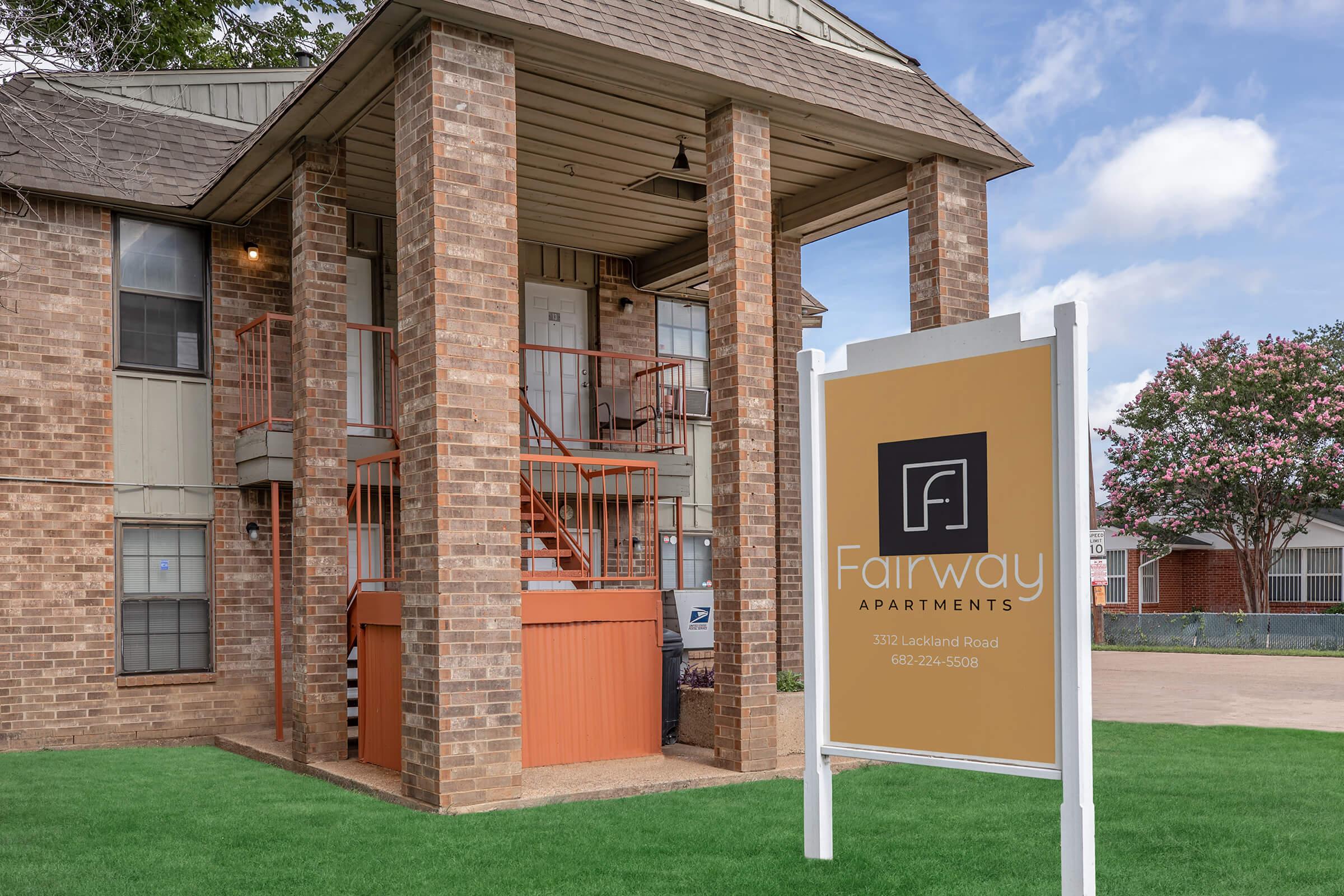 Exterior view of Fairway Apartments featuring a brick building and a large sign. The entrance includes a staircase leading to upper floors, and there are trees in the background with a partly cloudy sky. The sign displays the name and contact information for the apartments.
