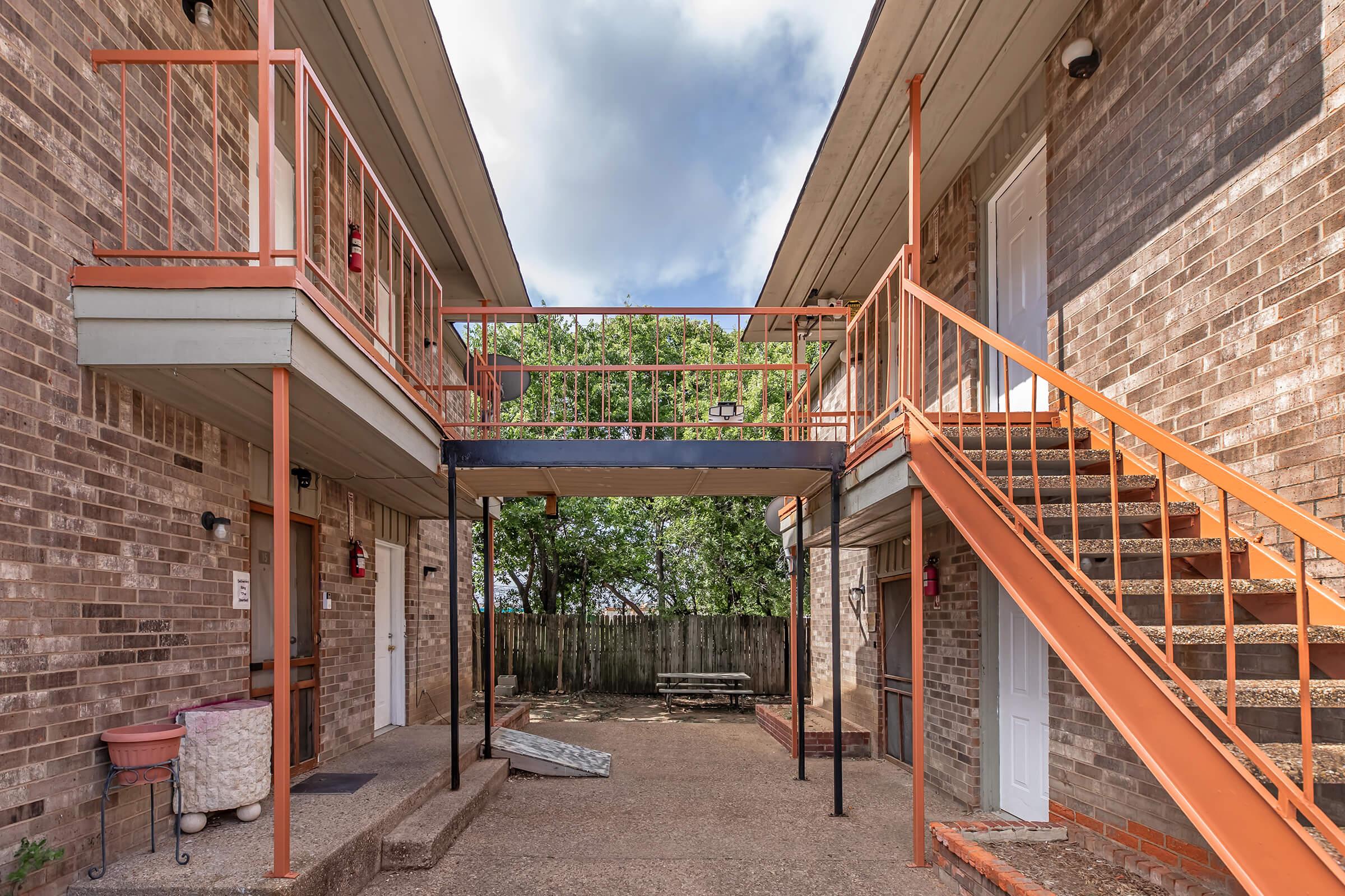 A view of a two-story apartment building with brick walls. There are orange railings and staircases leading to upper units. A picnic table is situated in a grassy area outside, surrounded by trees, under a partly cloudy sky.