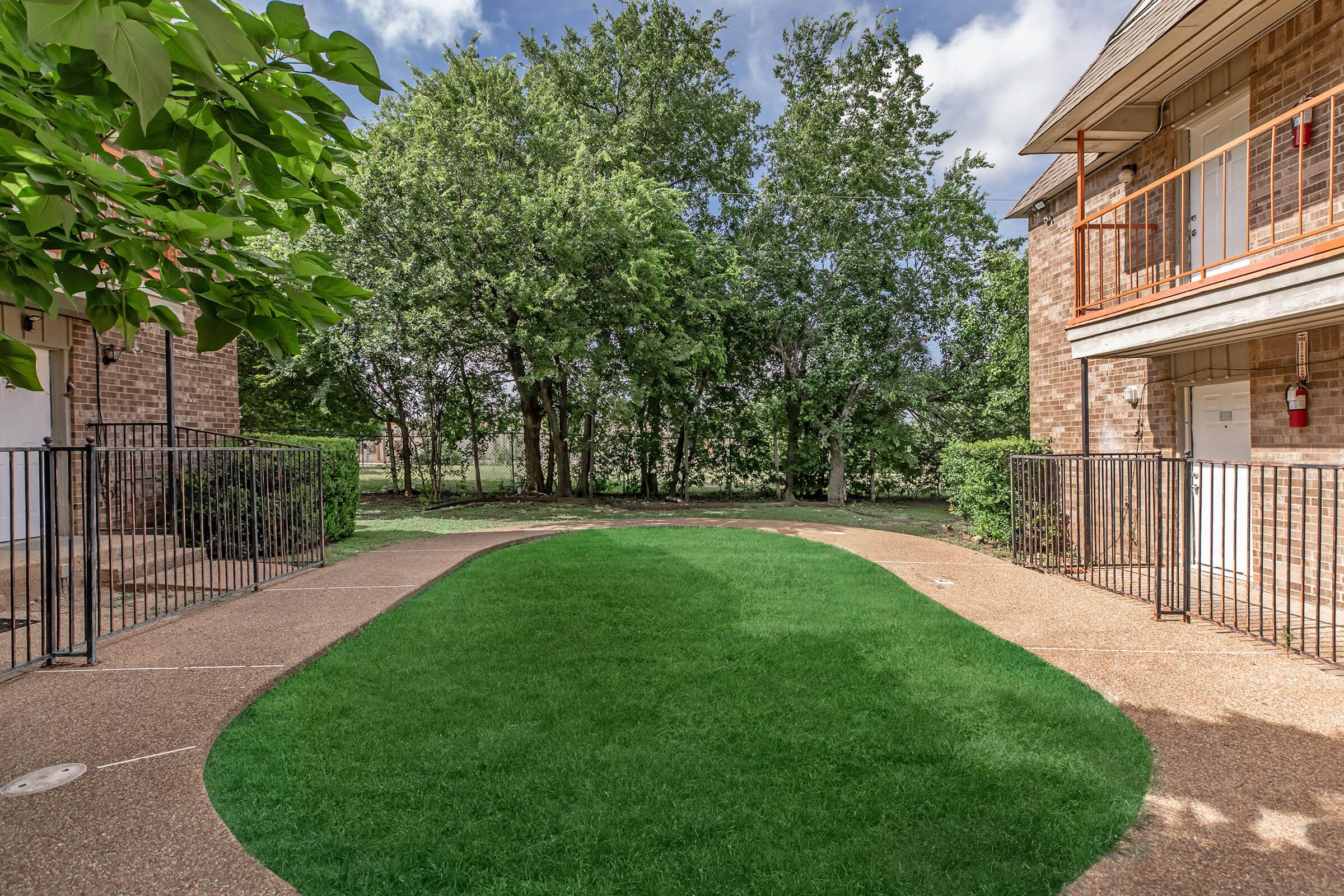 A well-maintained outdoor area featuring a lush green lawn bordered by a curved pathway. Surrounding trees provide shade, and there are two brick buildings on either side with balconies. A black fence encloses the space, creating a welcoming atmosphere for residents and visitors.