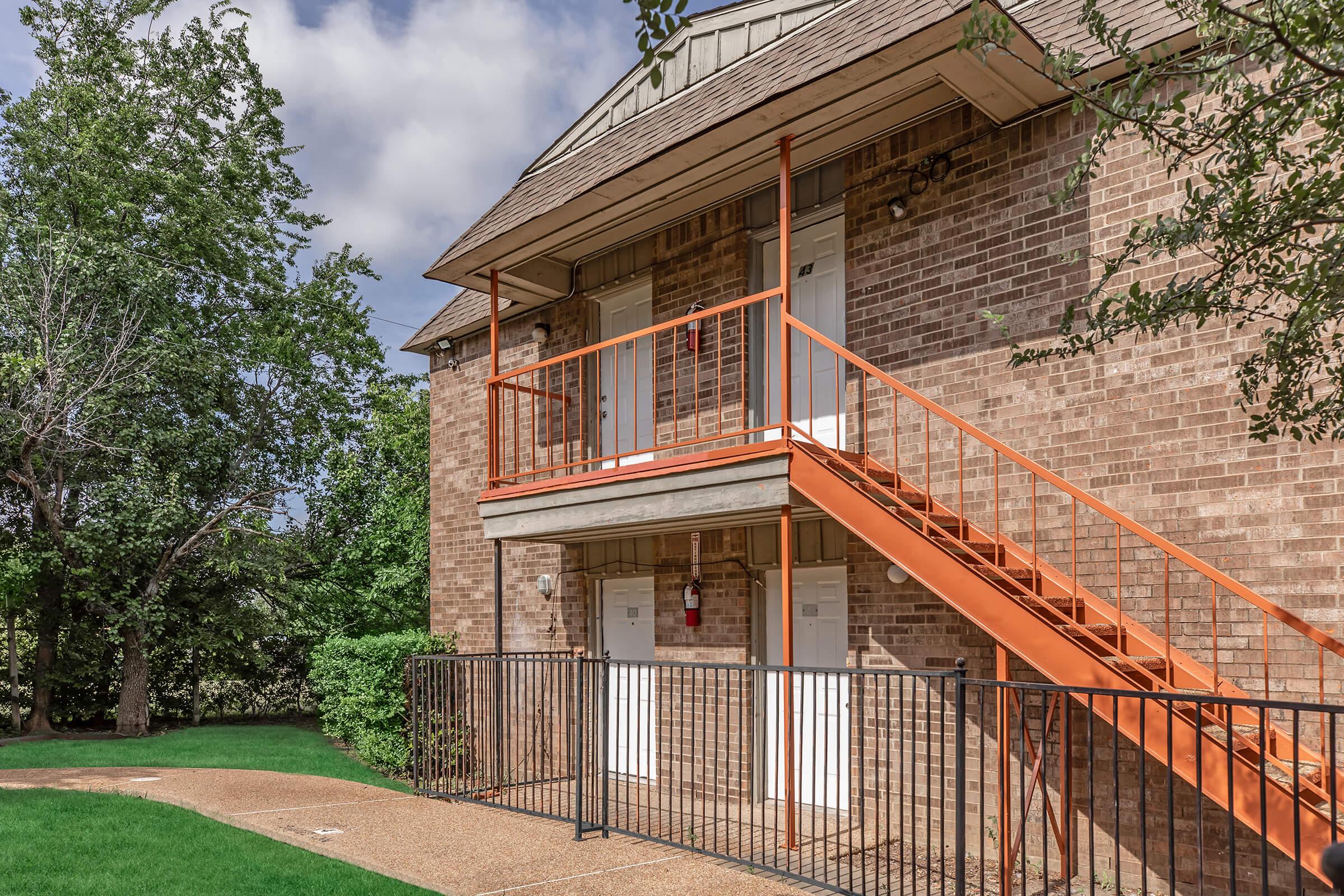 Exterior view of a two-story brick apartment building featuring a set of orange stairs leading to the second floor. The entrance doors are white, and there is a grassy area with a pathway in front. Surrounding greenery adds to the outdoor space, under a partly cloudy sky.