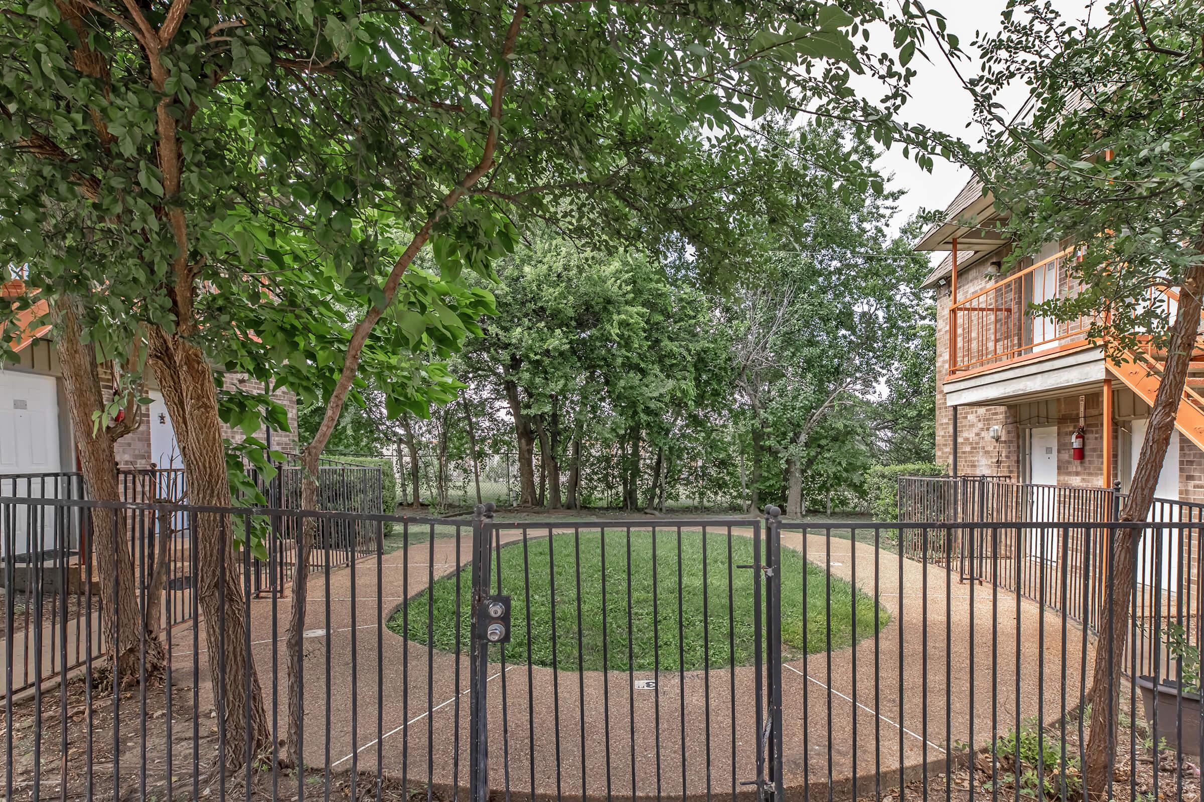 A fenced circular garden area with lush green grass surrounded by trees. In the background, there are two multi-story apartment buildings with orange accents. The scene is serene and inviting, showcasing a mix of natural and residential elements.