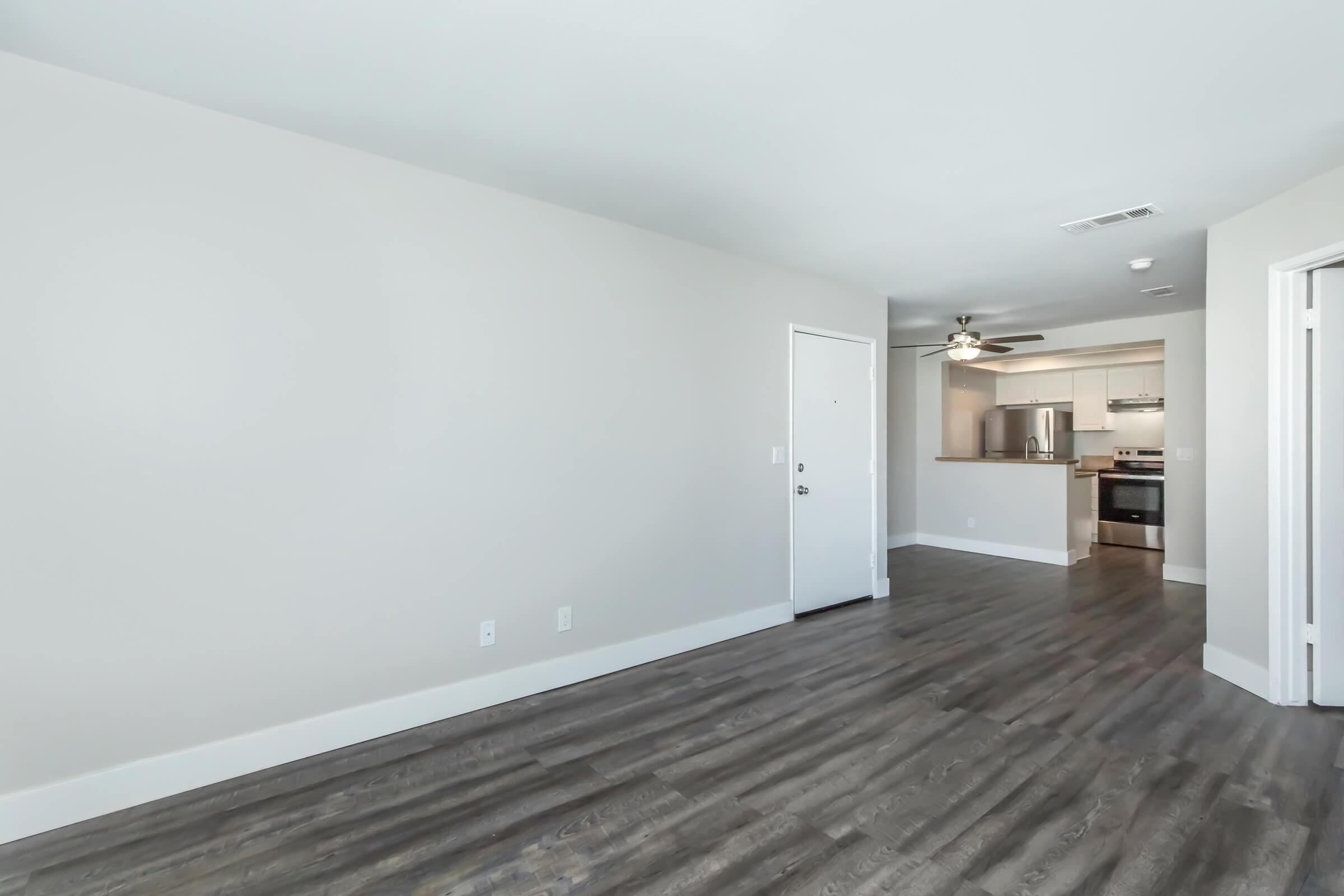A bright, modern interior of an apartment showcasing an empty living area with light-colored walls and dark wood flooring. A door leads outside, while an open kitchen area is visible in the background, featuring stainless steel appliances and a ceiling fan. The space is well-lit and inviting.