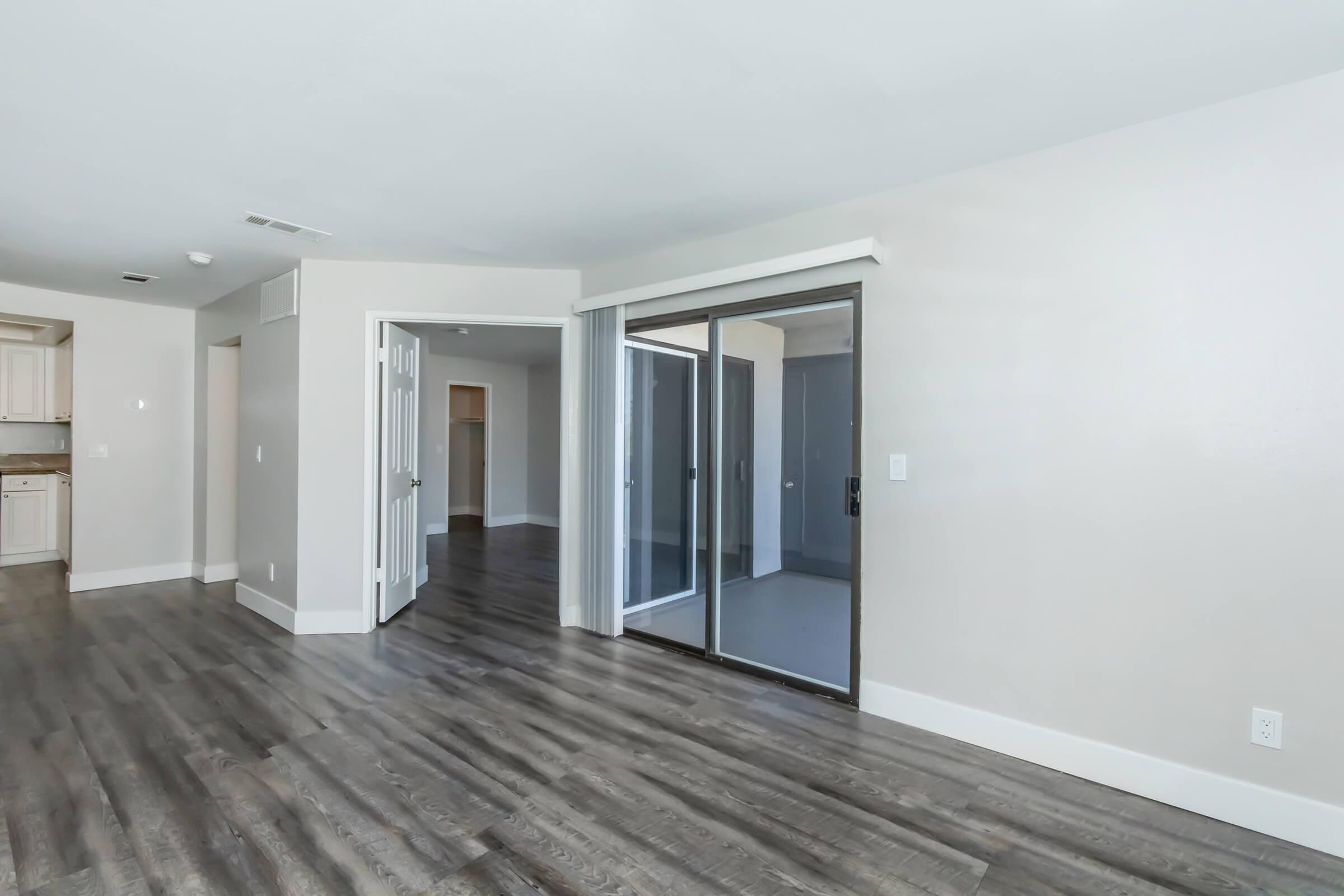 Interior view of an open living space showing hardwood flooring, a sliding glass door leading to an outdoor area, and neutral-colored walls. The kitchen is visible in the background, featuring light cabinetry. The room is well-lit and spacious, suitable for various furniture arrangements.