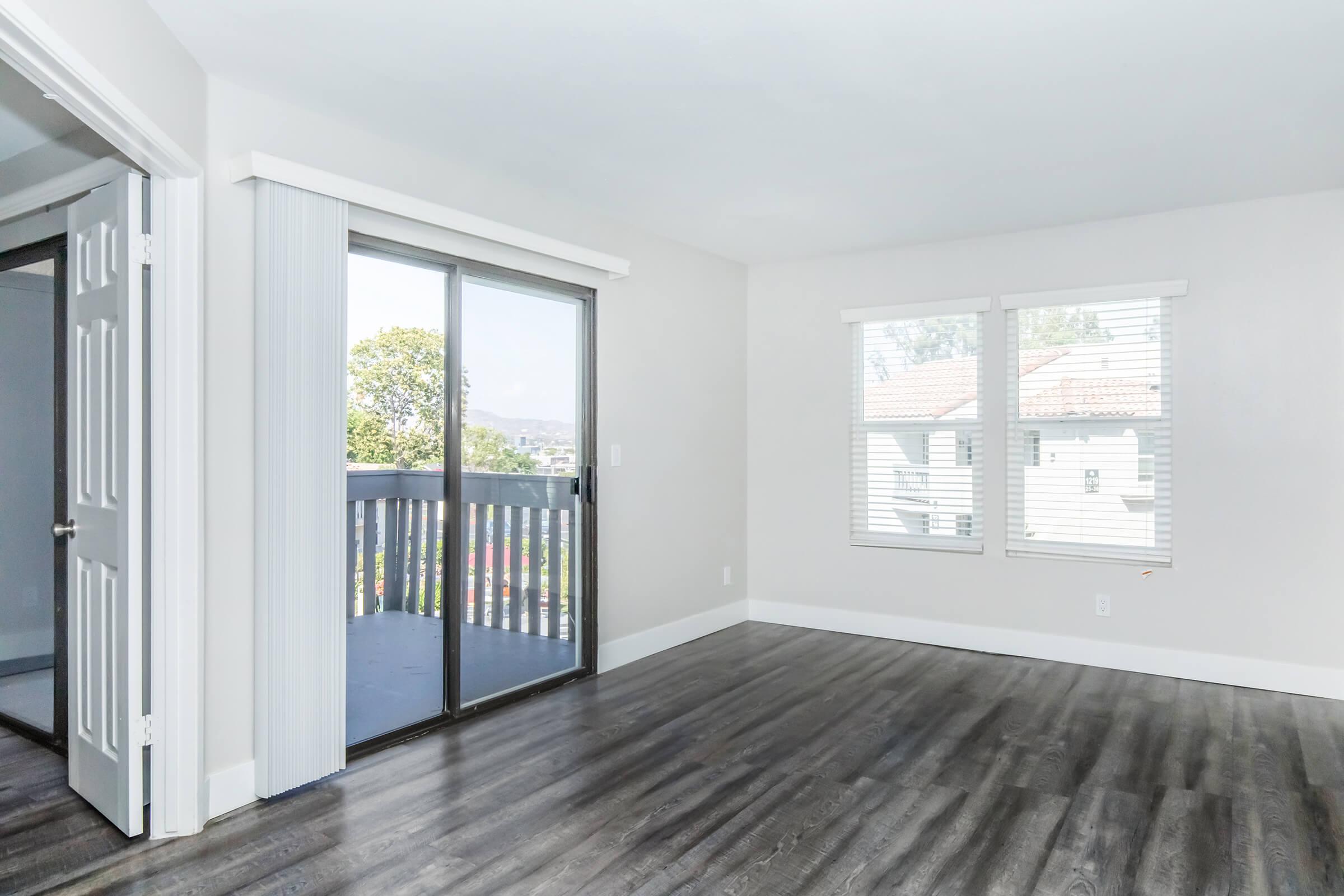 A bright and airy living room featuring laminate flooring, sliding glass doors leading to a balcony, and two windows with white blinds. The walls are painted in a light color, and there is a door leading to another room. The space is open and uncluttered, creating a welcoming atmosphere.