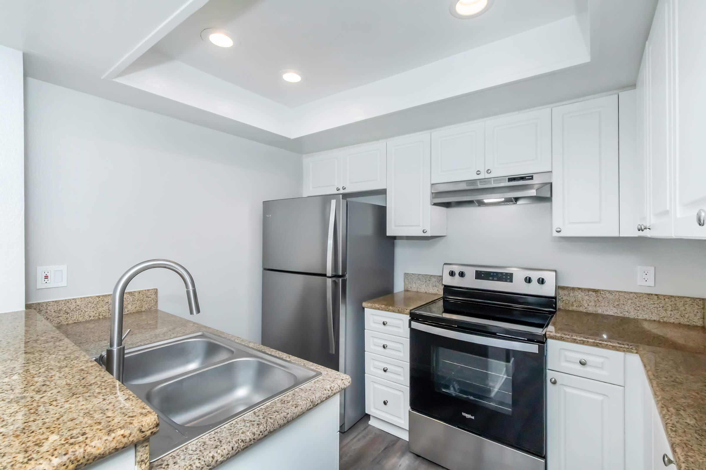 A modern kitchen featuring white cabinetry, a stainless steel refrigerator, an oven, and a granite countertop. The space is well-lit with recessed lighting and has a double sink with a sleek faucet. The flooring is a dark wood laminate.