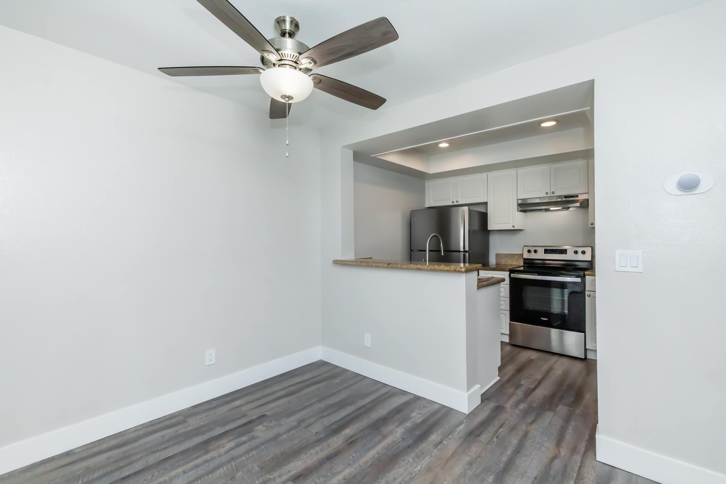 Modern kitchen and living area featuring a ceiling fan, granite countertop, stainless steel appliances, and light gray walls. The open layout connects the kitchen to the living space, showcasing a clean and inviting atmosphere with wood-like flooring.