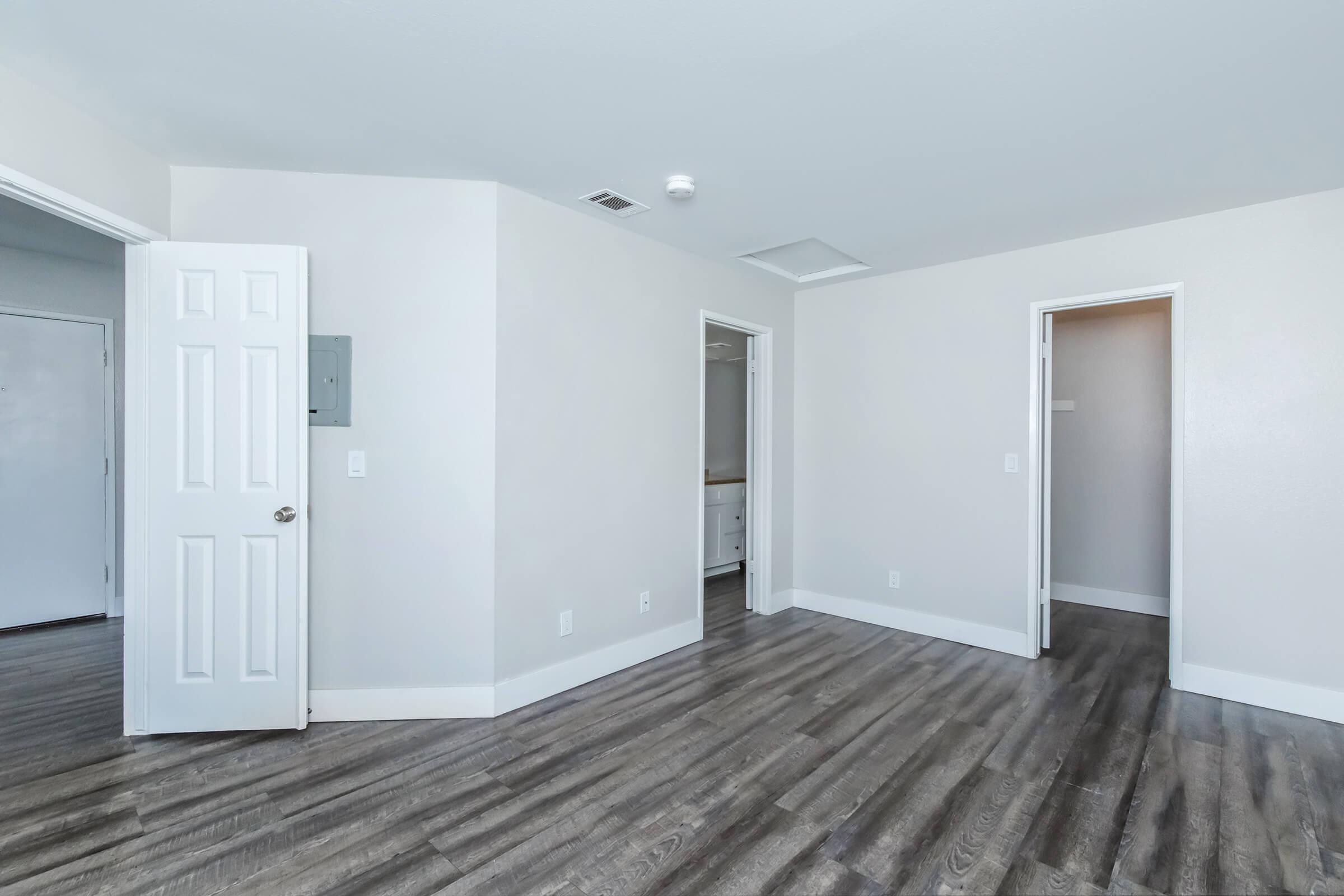 Empty room with light gray walls and wooden laminate flooring. There are two doorways leading to other spaces, and a utility panel is visible on the left wall. The room is well-lit, creating a spacious and open feel.