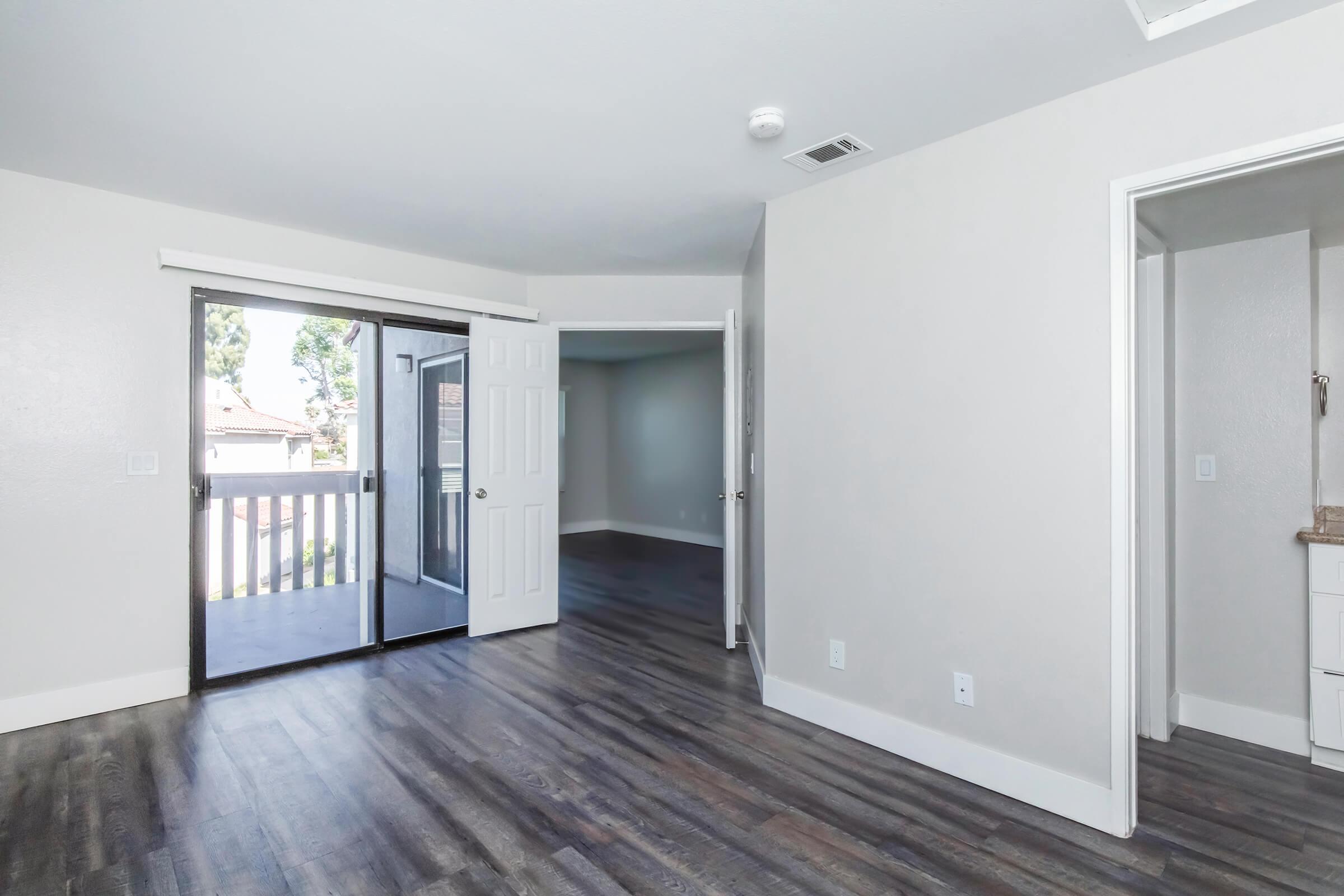 Interior of a modern room featuring a sliding glass door that opens to a balcony, light gray walls, and new wood-style flooring. A doorway leads to another room, and a small bathroom with a sink is visible on the right side. The space is well-lit with plenty of natural light.