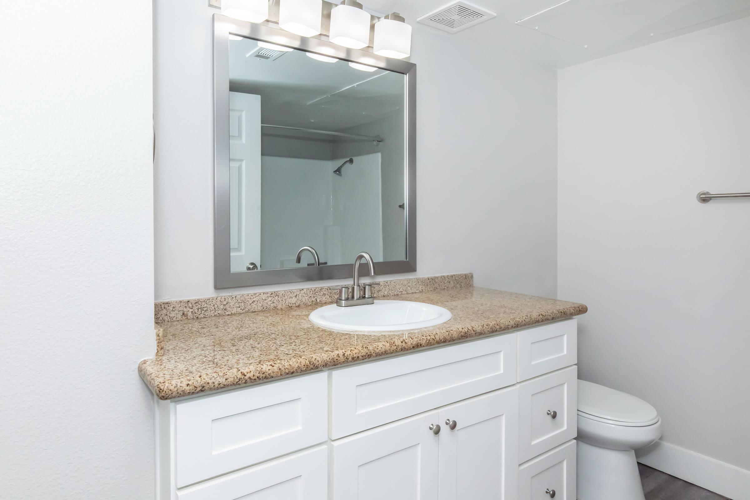 A modern bathroom featuring a white cabinet with a granite countertop and a round sink. Above the sink is a rectangular mirror with lights, and a toilet is visible nearby. The walls are painted in light gray, and there's a shower area partially visible in the background. The overall design is clean and contemporary.