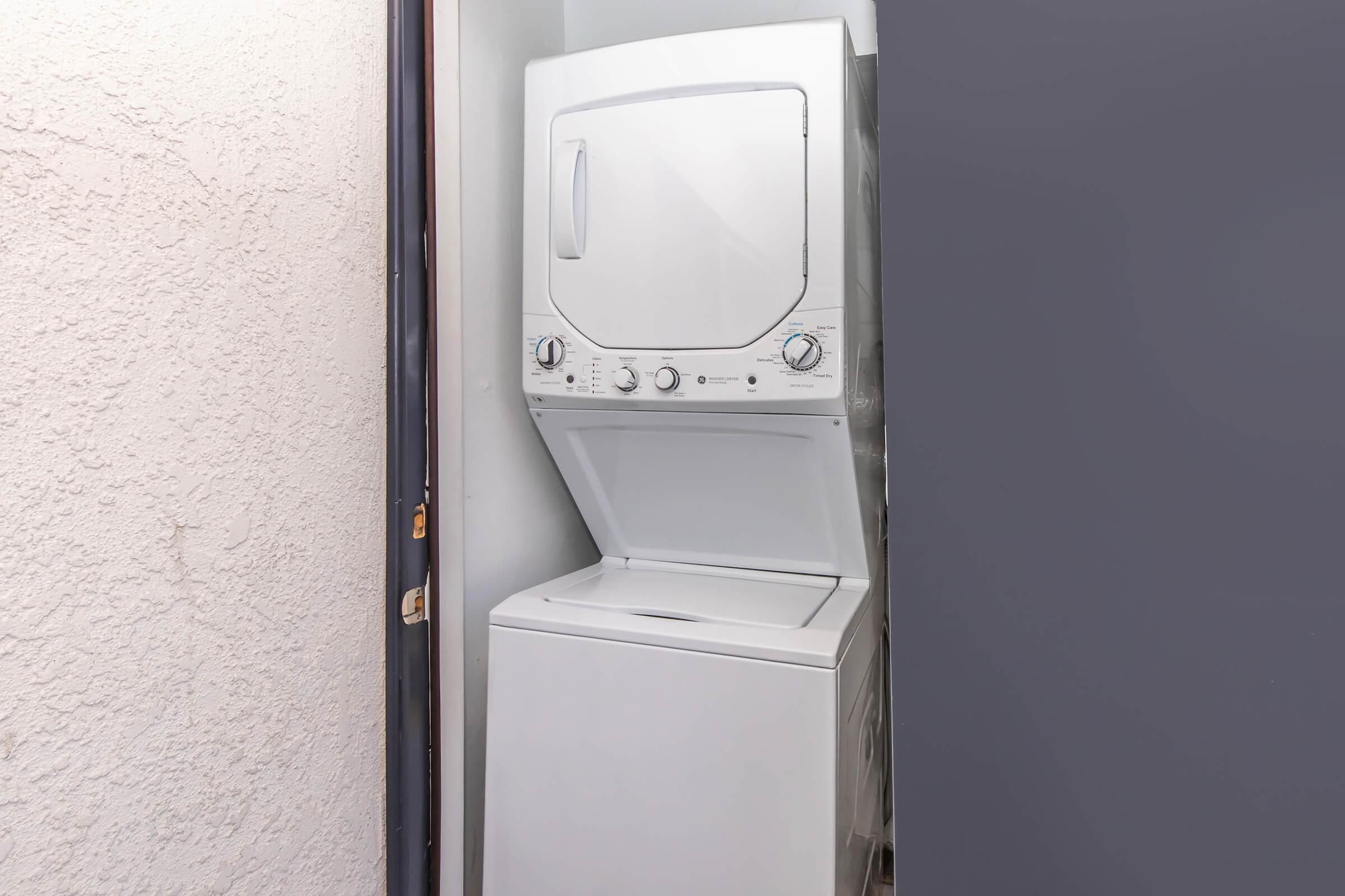 A stackable washer and dryer unit positioned in a narrow laundry space, with a textured wall in the background and a closed door frame on the left. The appliances feature control knobs and a clean, modern design.