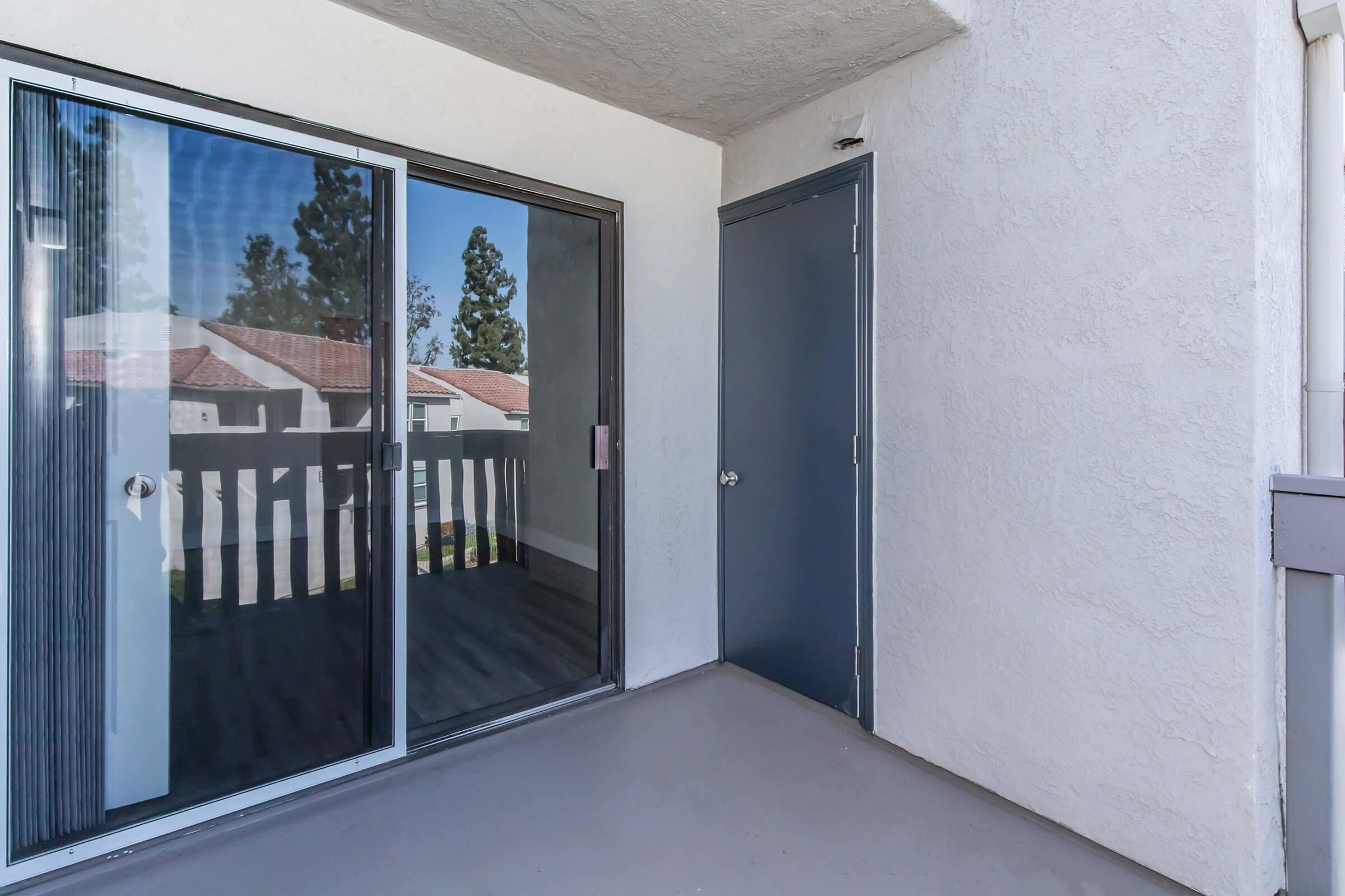 A small balcony with sliding glass doors leading to the interior. The floor is painted gray, and a closed door is visible on one side. The surrounding area includes trees and a view of nearby buildings. The walls are light-colored, creating a bright and open space.