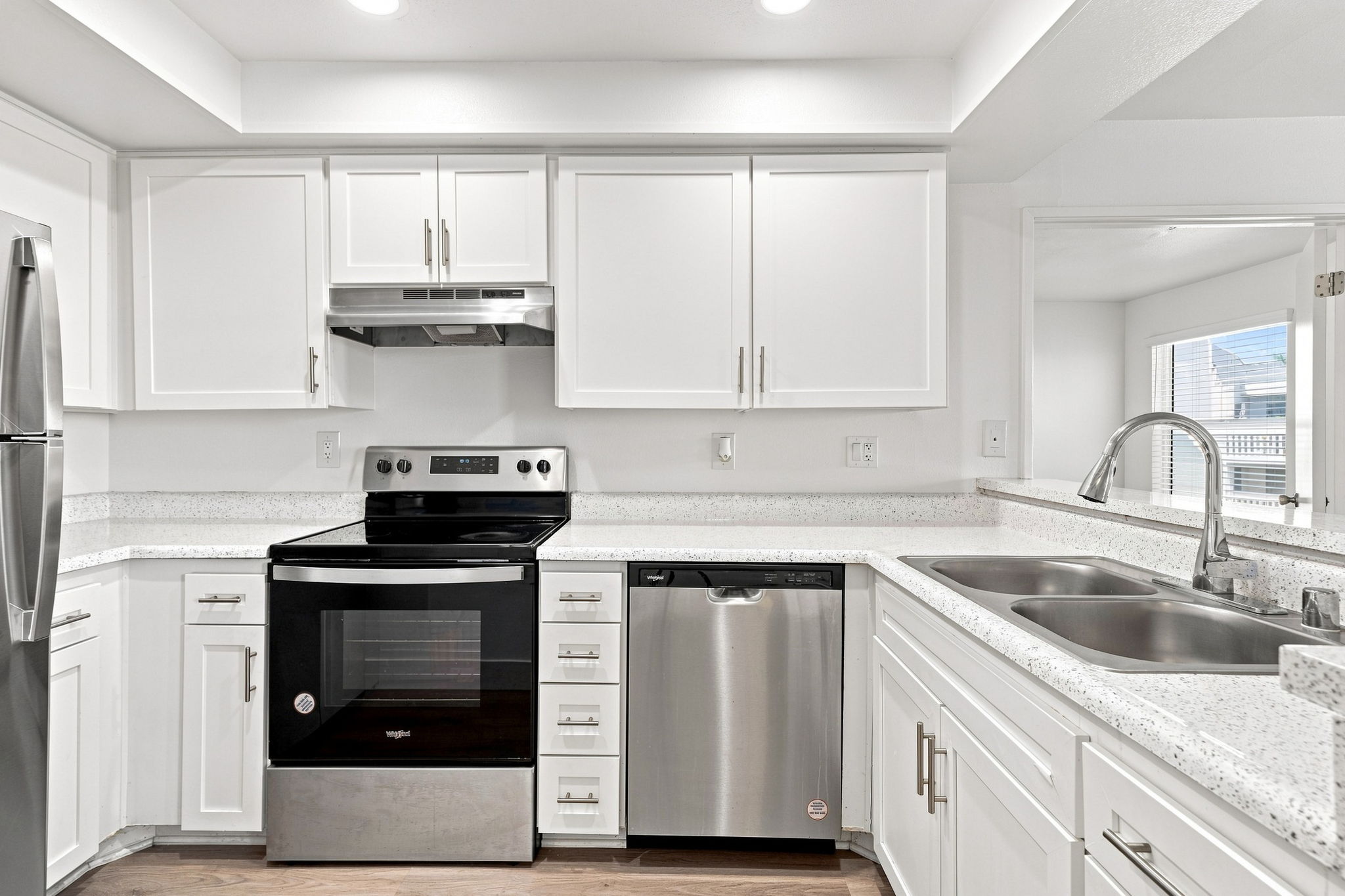 A modern kitchen featuring white cabinets and countertops, a stainless steel refrigerator, an electric stove with an oven, a dishwasher, and double sink. Bright lighting enhances the spacious layout, highlighting the clean lines and contemporary design.