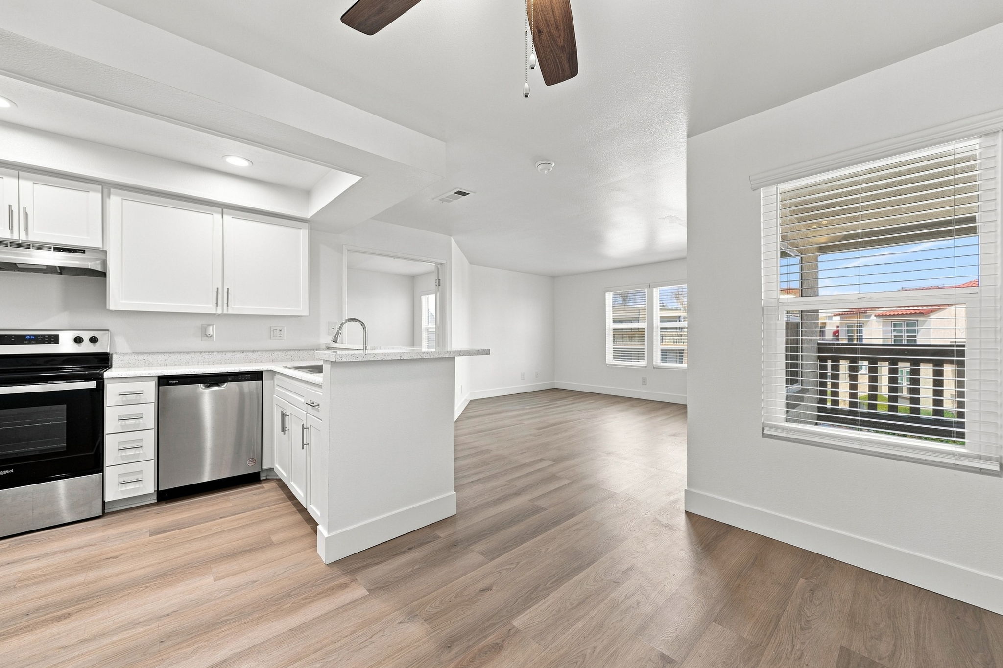 A modern, open-concept kitchen and living area featuring white cabinetry, stainless steel appliances, and a ceiling fan. The kitchen includes a breakfast bar and is connected to a spacious living room with large windows. Light-colored wood flooring runs throughout the space, enhancing the bright and airy atmosphere.