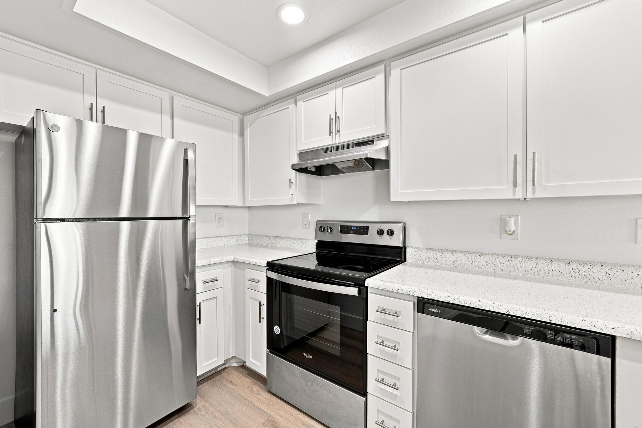 Modern kitchen featuring stainless steel appliances including a refrigerator, oven, and dishwasher. The cabinetry is white with sleek handles, and the countertop has a speckled design. Bright overhead lighting enhances the clean, contemporary look of the space.