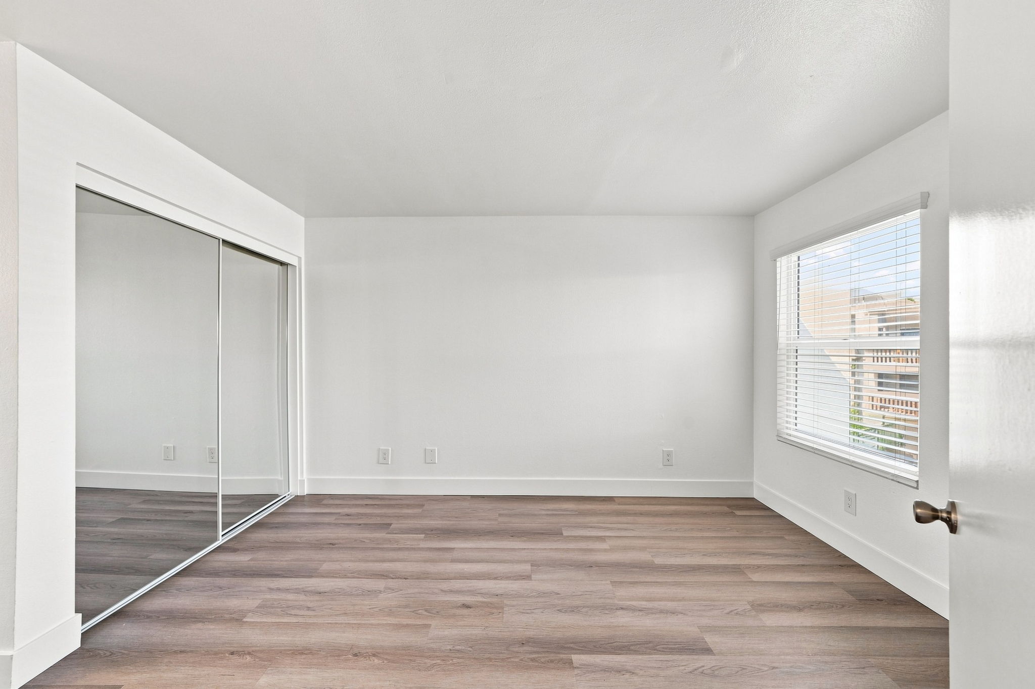 A bright, empty room featuring light-colored walls and a large window with blinds. There is a mirrored closet on one side, and the flooring is made of light wood. Natural light fills the space, creating an open and airy atmosphere.