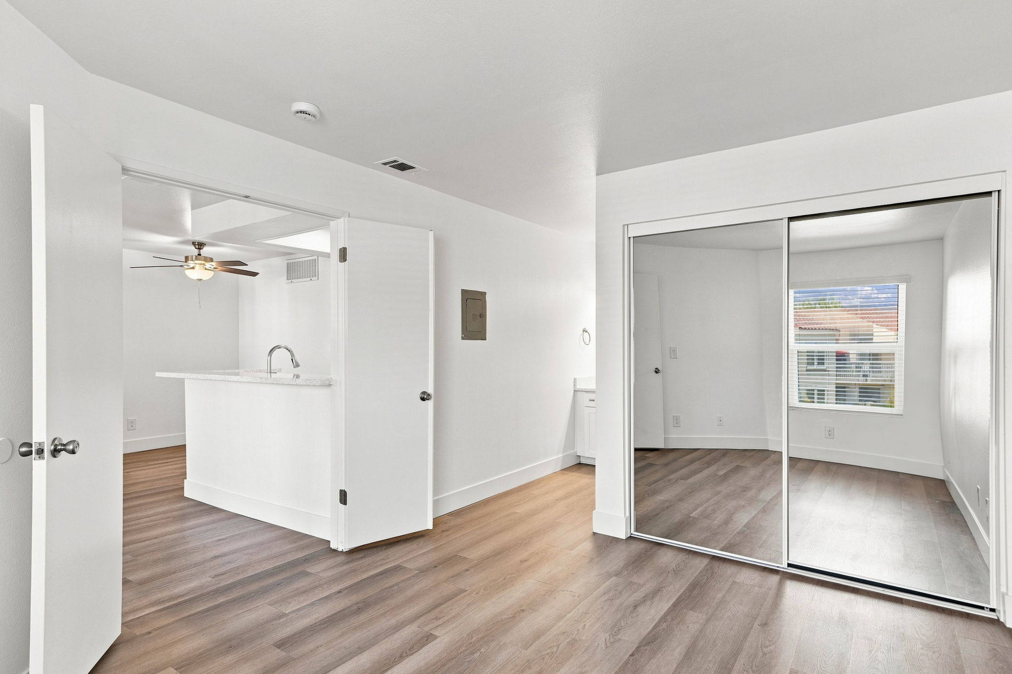 A bright, airy interior of a room featuring white walls, a ceiling fan, and laminate flooring. On one side, there is a mirrored closet door, and on the opposite side, an open doorway leads to a kitchen area with a smooth countertop. Natural light streams in through a window, creating a welcoming atmosphere.