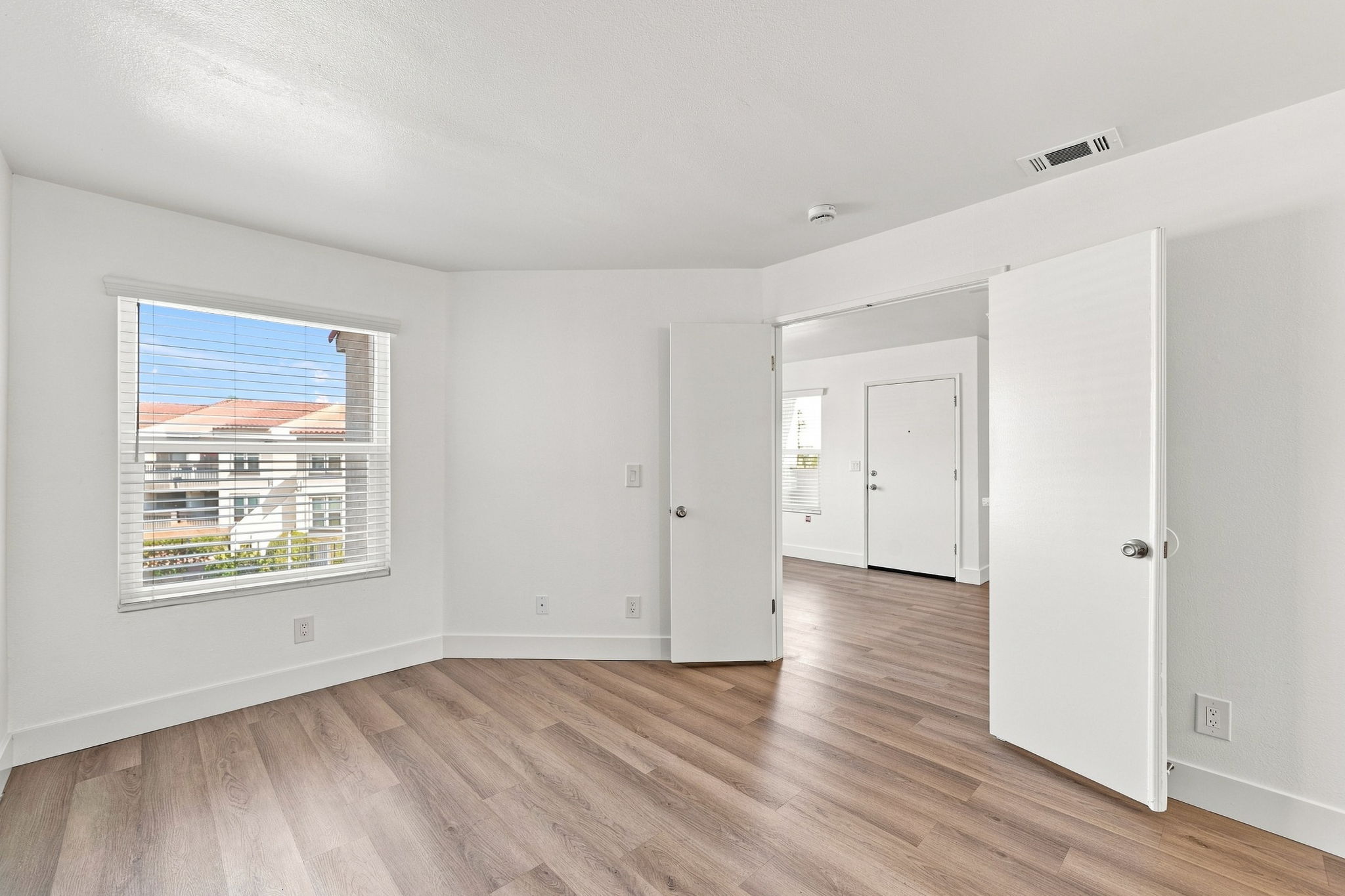 Bright, empty interior of a modern room featuring light-colored walls, wooden flooring, and multiple doorways. A window with blinds allows natural light to enter, showcasing a view of the outdoors. The space is clean and minimalist, ideal for various uses or personal decoration.