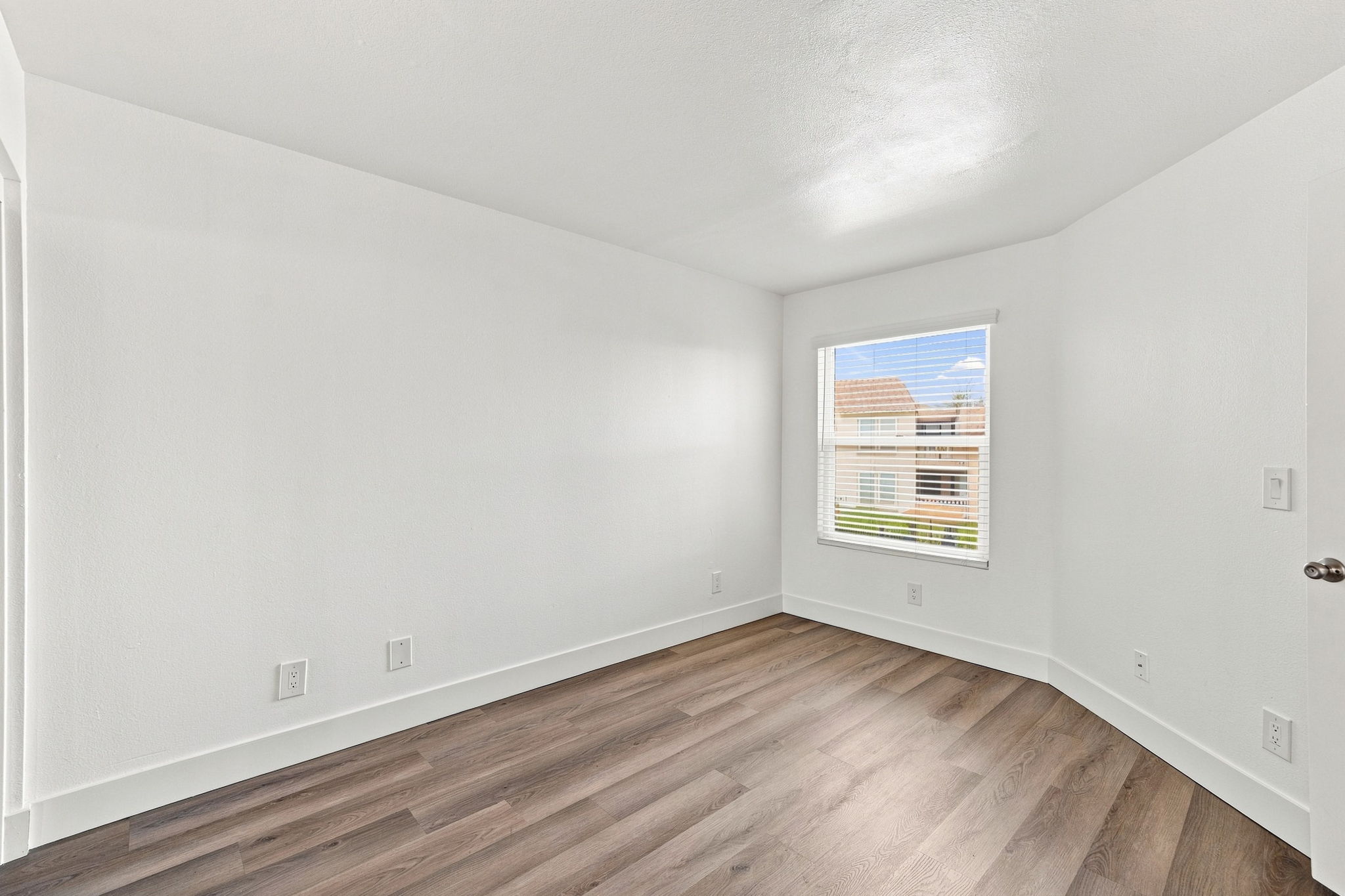 A bright, empty room with white walls and a window. The flooring is dark wood laminate, and there are no furnishings or decorations. Natural light is coming through the window, illuminating the space.