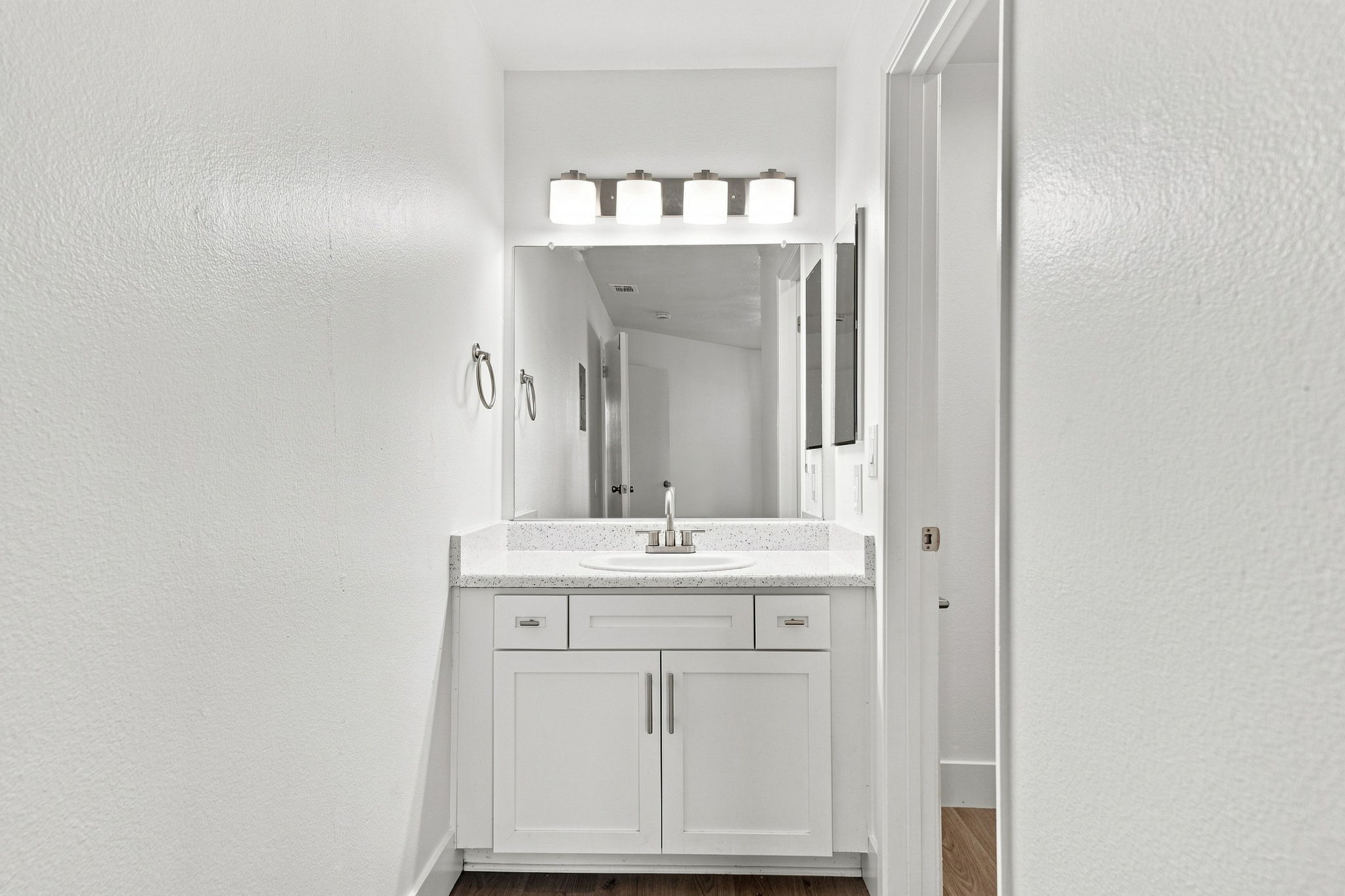 A clean, modern bathroom featuring a white vanity with a sink, a large mirror above it, and four light fixtures. The walls are painted white, and there are two towel holders on either side of the vanity. The floor has a warm wood finish, and there’s a glimpse of a doorway on the left.