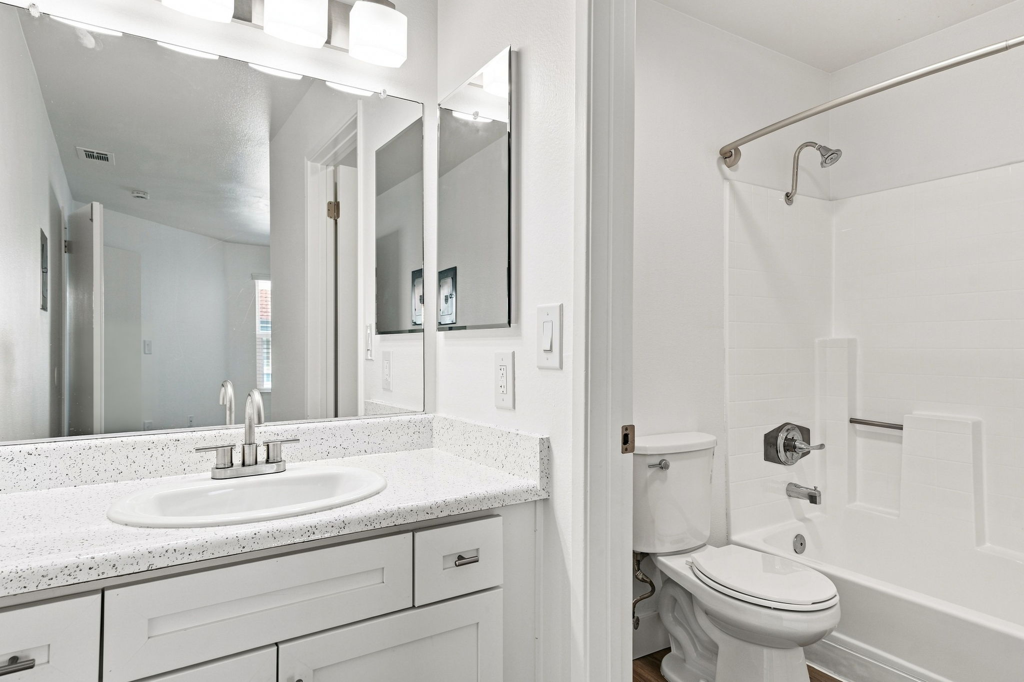 A modern bathroom featuring a white countertop with a sink and two faucets, a large mirror above, a shower-tub combo with a silver shower rod, a toilet, and light-colored walls. Natural light enters from a window in the background, creating a bright and clean atmosphere.
