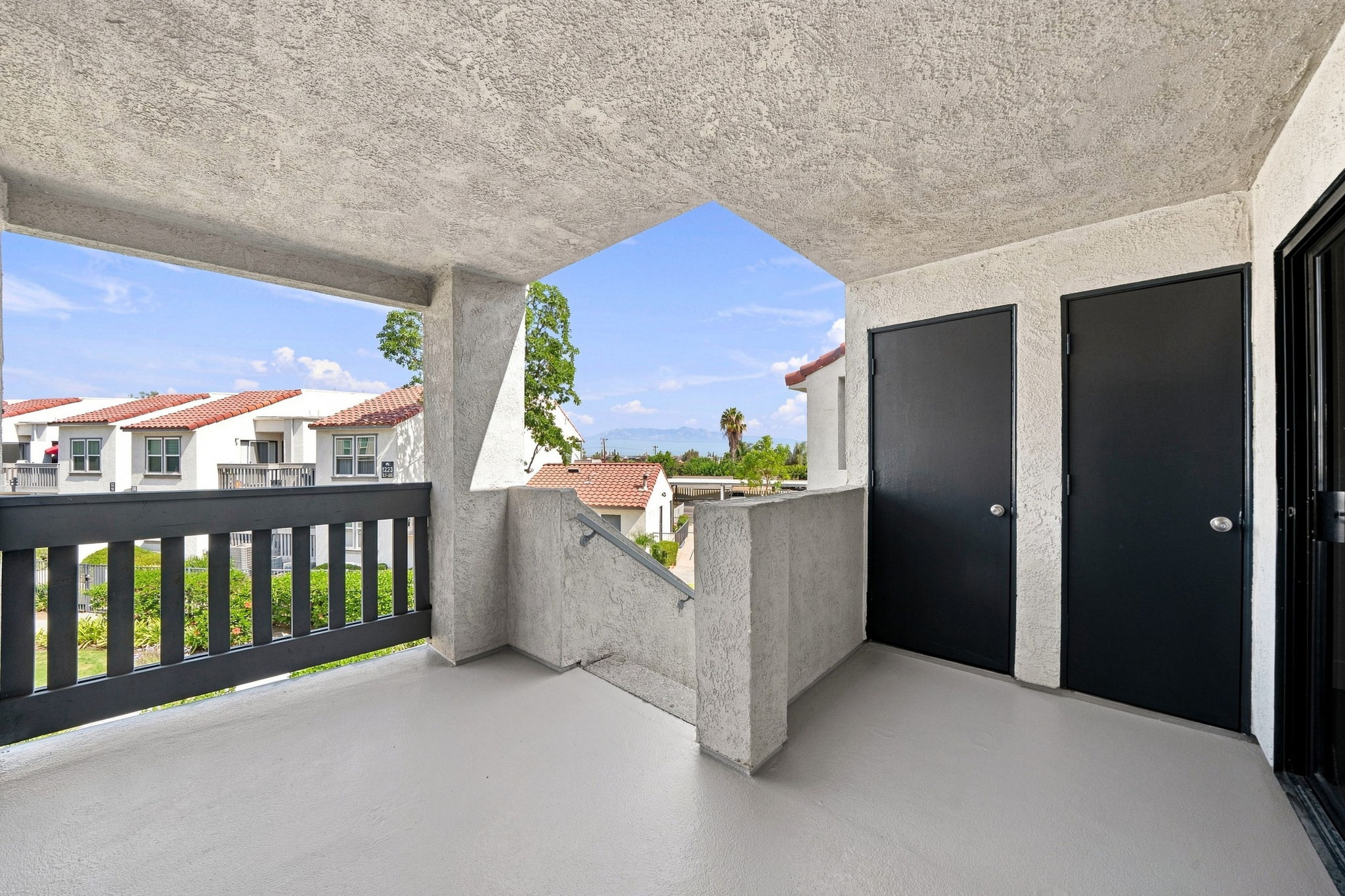 A view from a balcony featuring a textured gray floor, black doors leading to adjacent rooms, and an open space with a scenic background of rooftops and greenery under a clear blue sky. The balcony has a unique geometric design with a partially visible staircase.