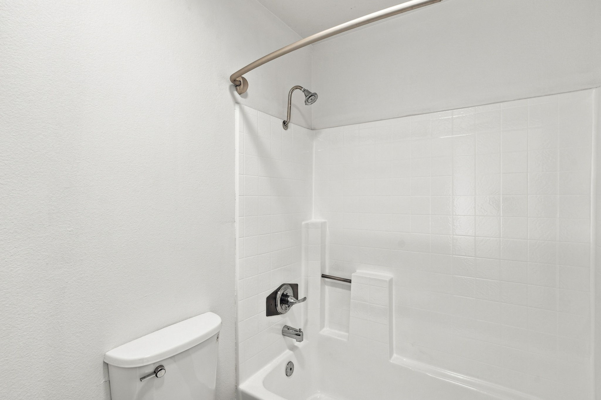 A clean, minimalist bathroom featuring a white bathtub with a showerhead, a curved shower rod, and a white toilet. The walls and tiles are predominantly white, creating a bright and spacious atmosphere. The design emphasizes functionality with an absence of decorative elements.